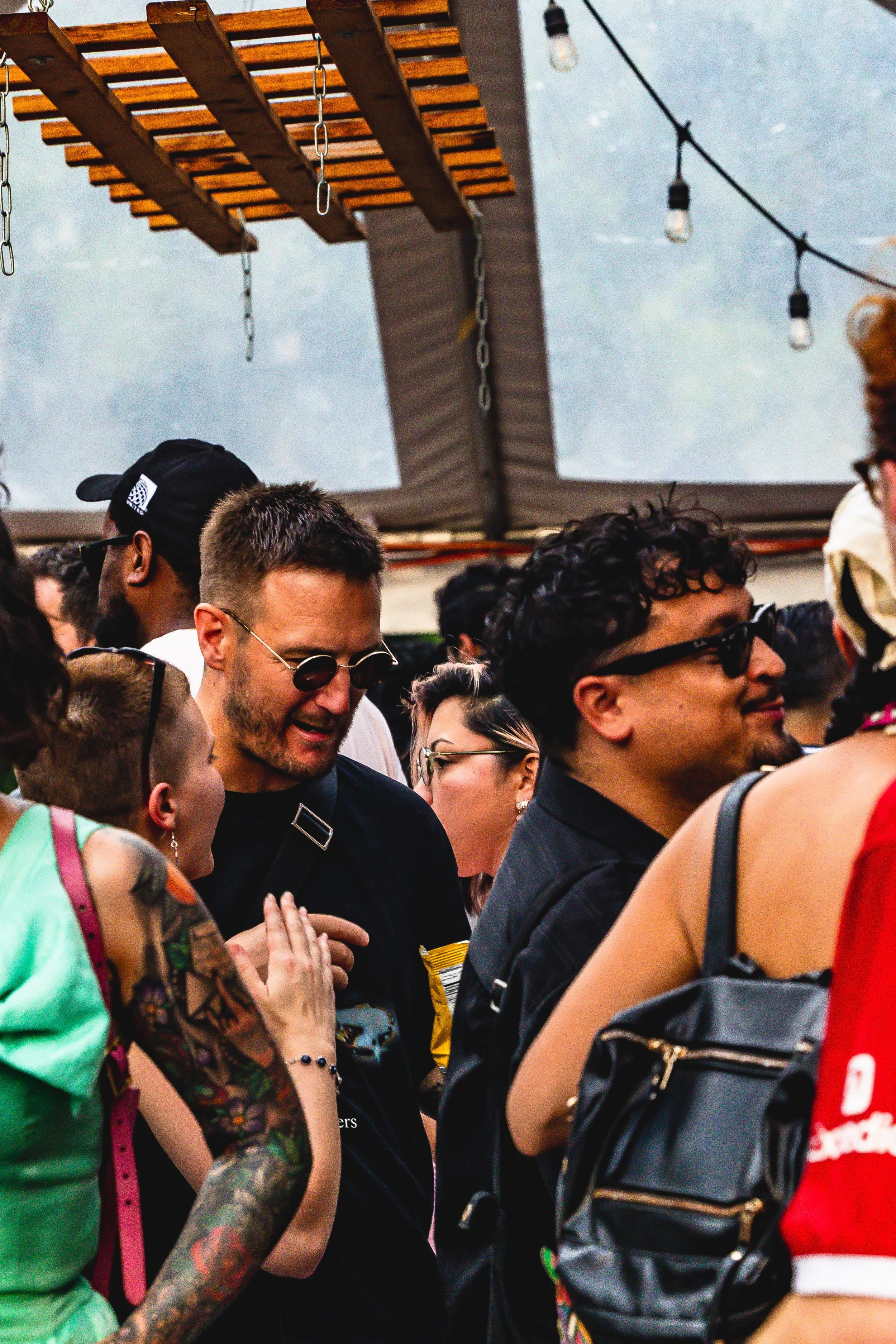 Crowd of diverse young adults socializing at an outdoor event or festival, under string lights and canopy roof, with various styles of clothing and accessories.