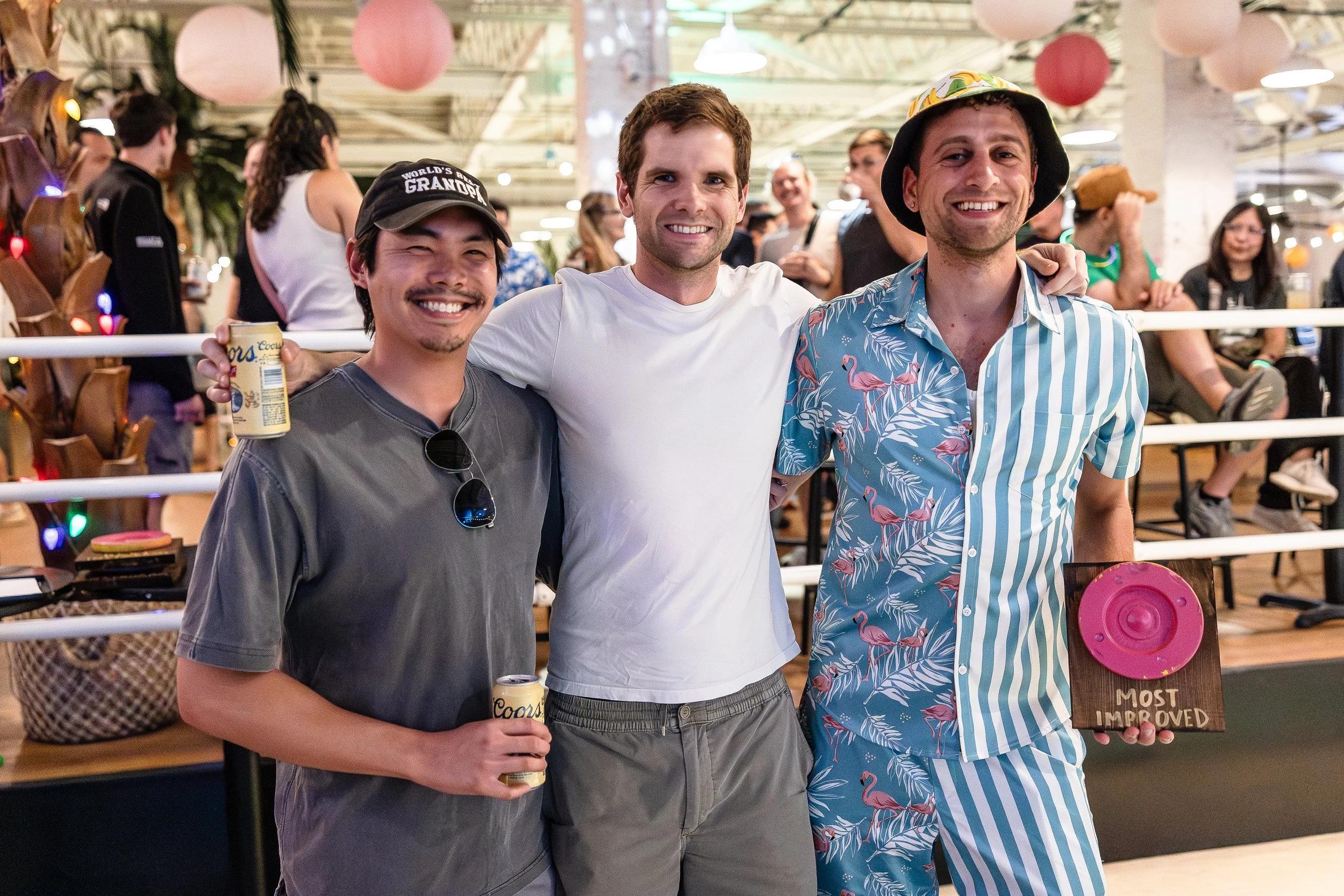 Three men standing together and smiling at a social gathering, two holding drinks, one holding a plaque that reads 'Most Improved', in a lively indoor setting with other people in the background.