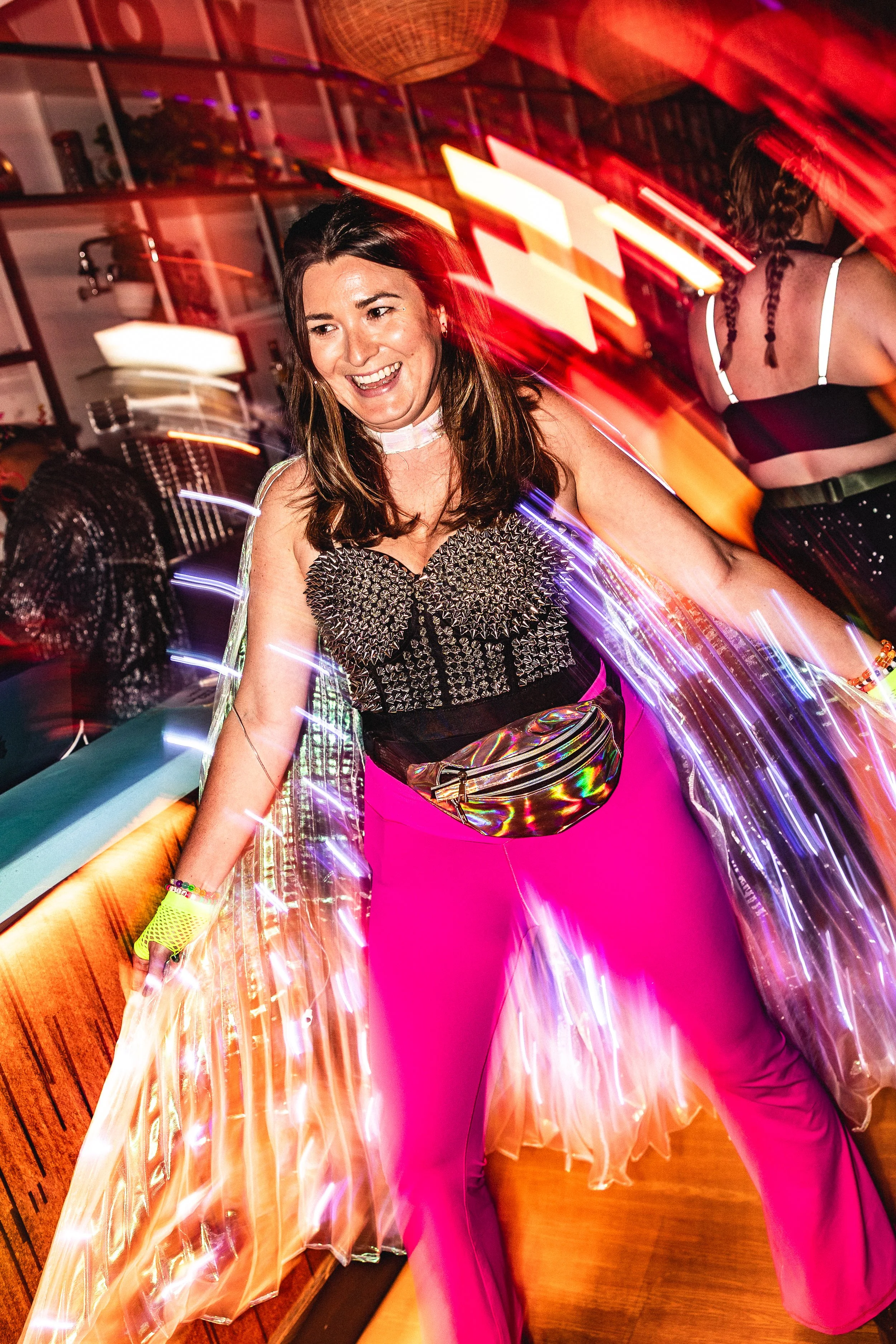 A woman dancing at a party or nightclub, wearing bright pink pants and a sparkly black top, with colorful light streaks surrounding her.