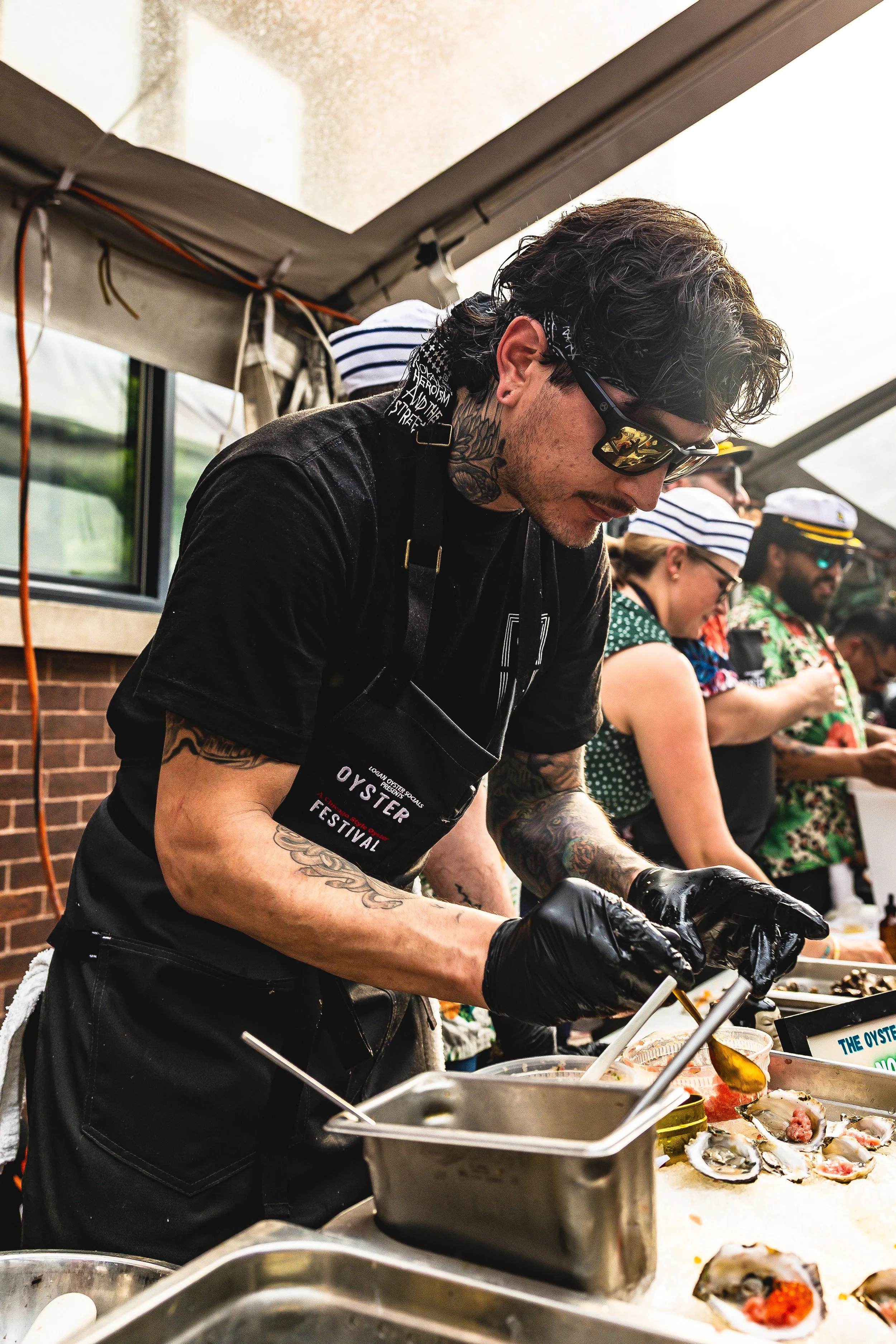 A man with tattoos, black gloves, and sunglasses opening oysters at a food festival, with others preparing food in the background under a tent.