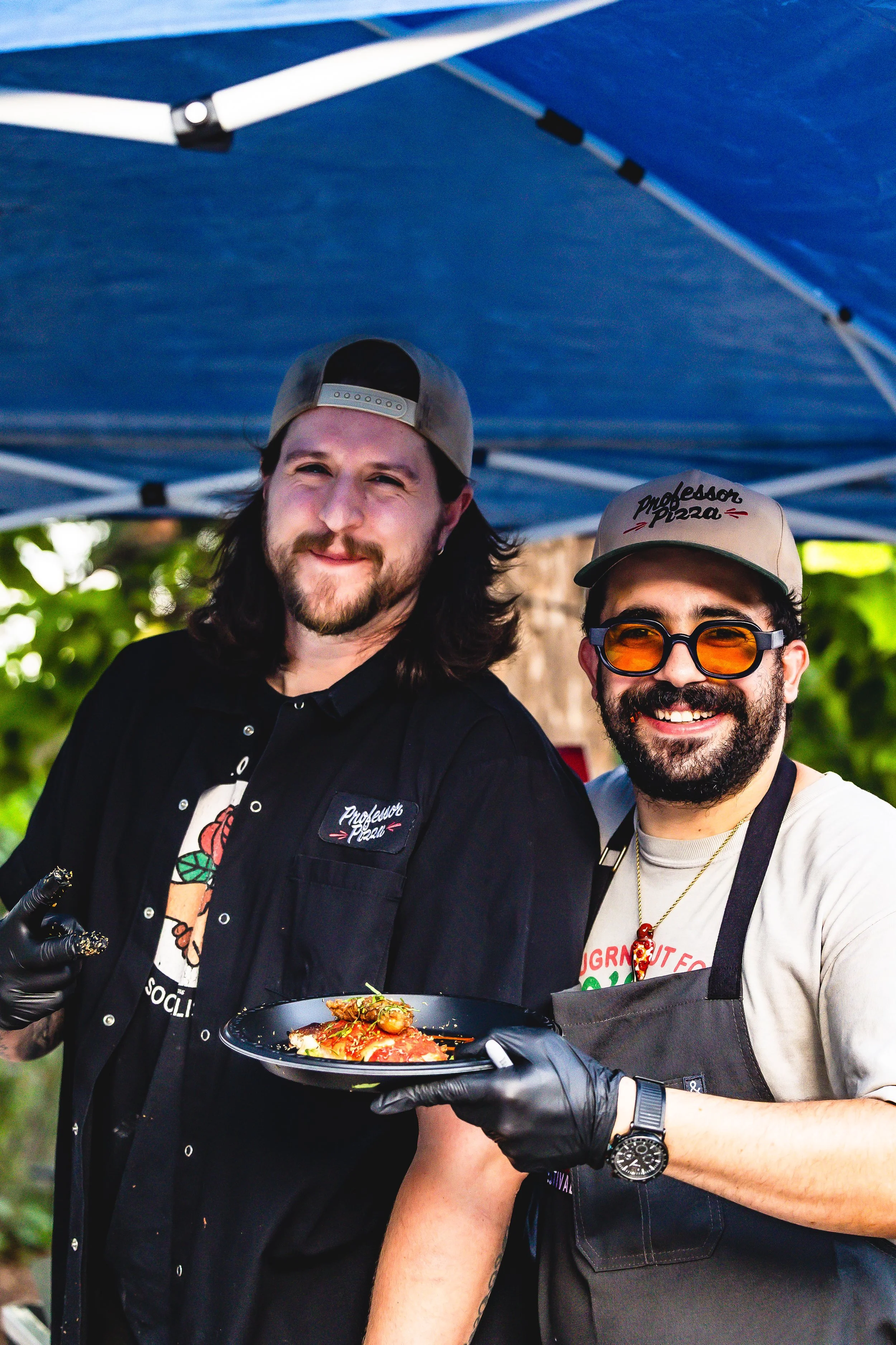 Two smiling men wearing caps and gloves, one holding a plate of food, standing outdoors under a blue canopy with green trees in the background.