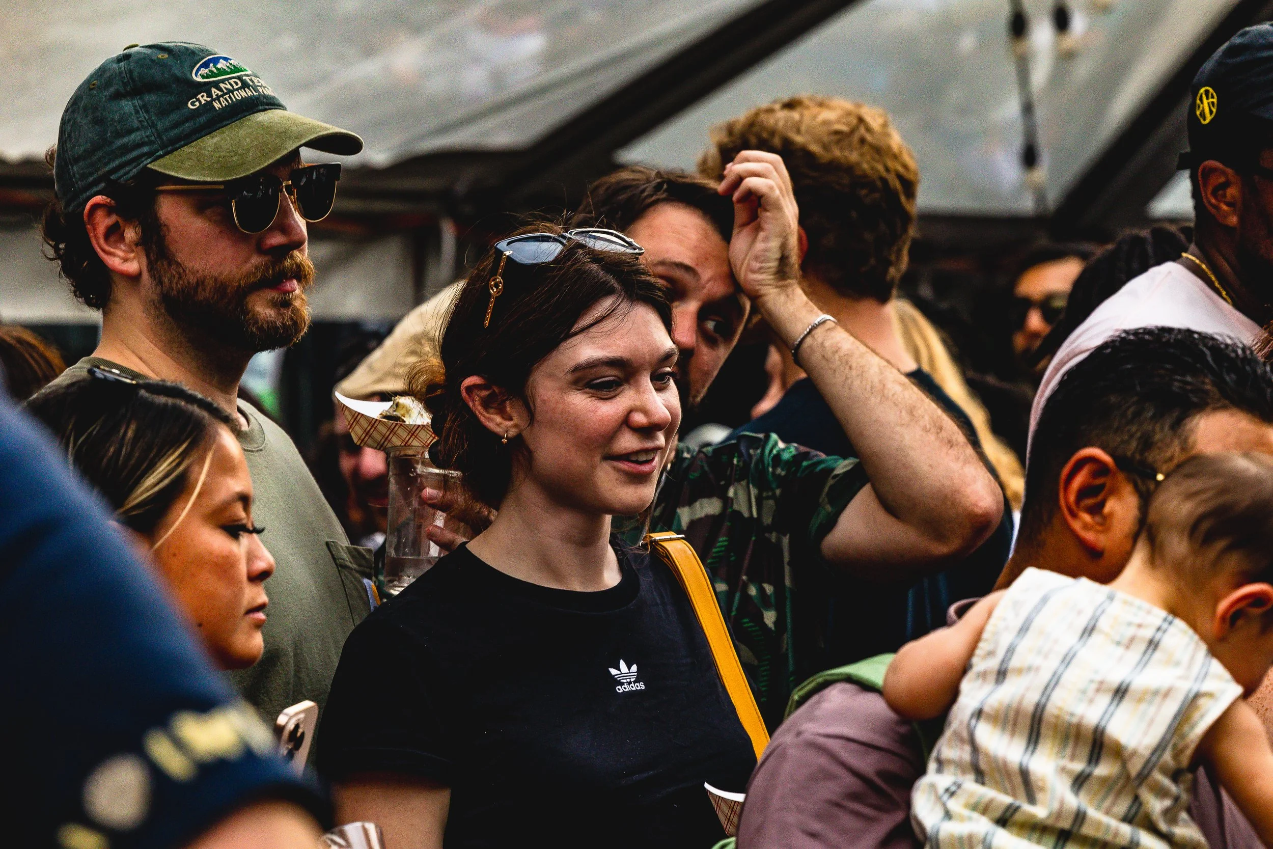 A group of diverse people at a crowded indoor event, some with glasses and casual clothing, some with children, engaged and observing something.
