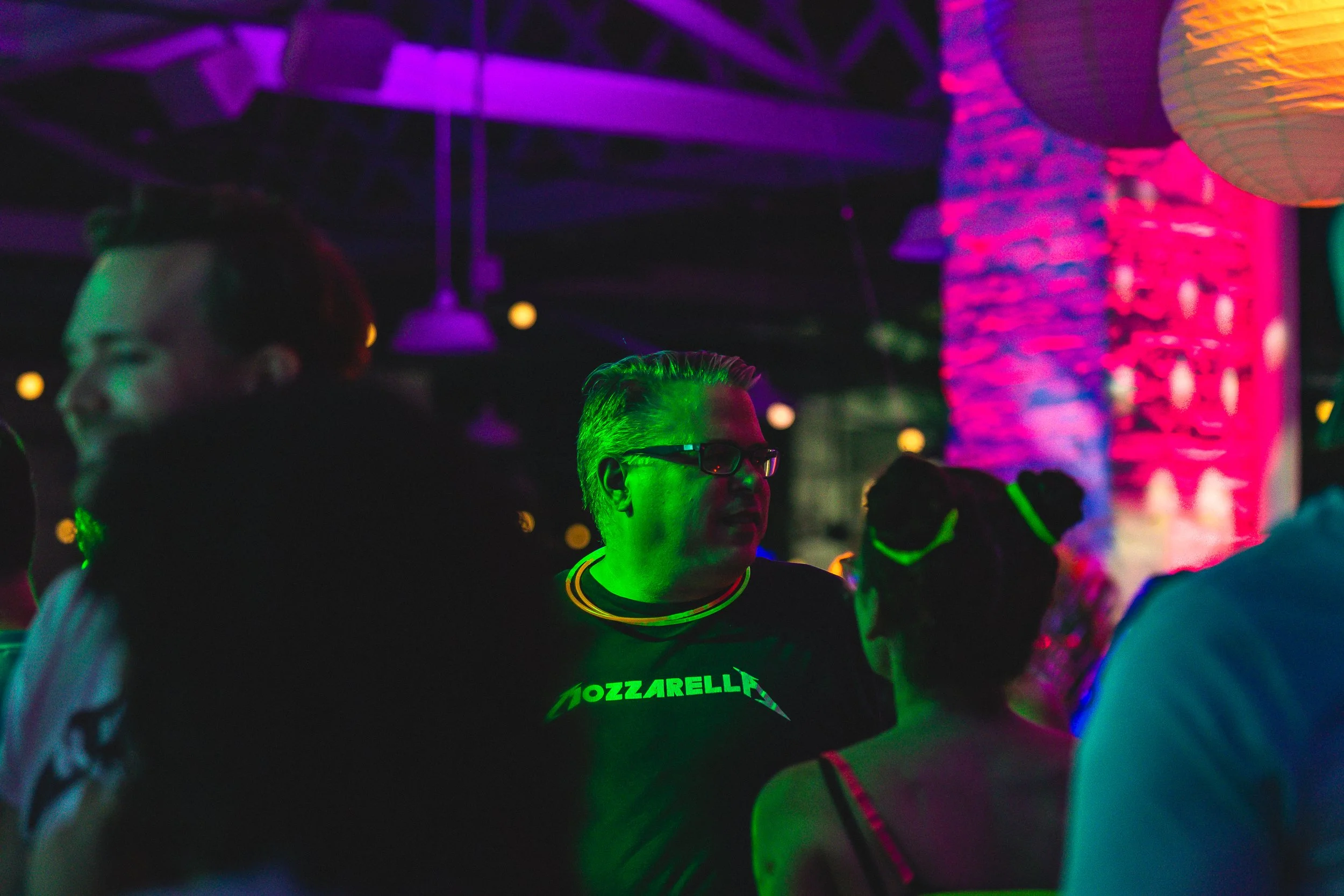 People socializing at a nightclub with colorful neon lighting and lanterns, including a man wearing glasses and a black T-shirt with 'Mozzarella' written on it, and a woman with glasses and a top with thin straps.
