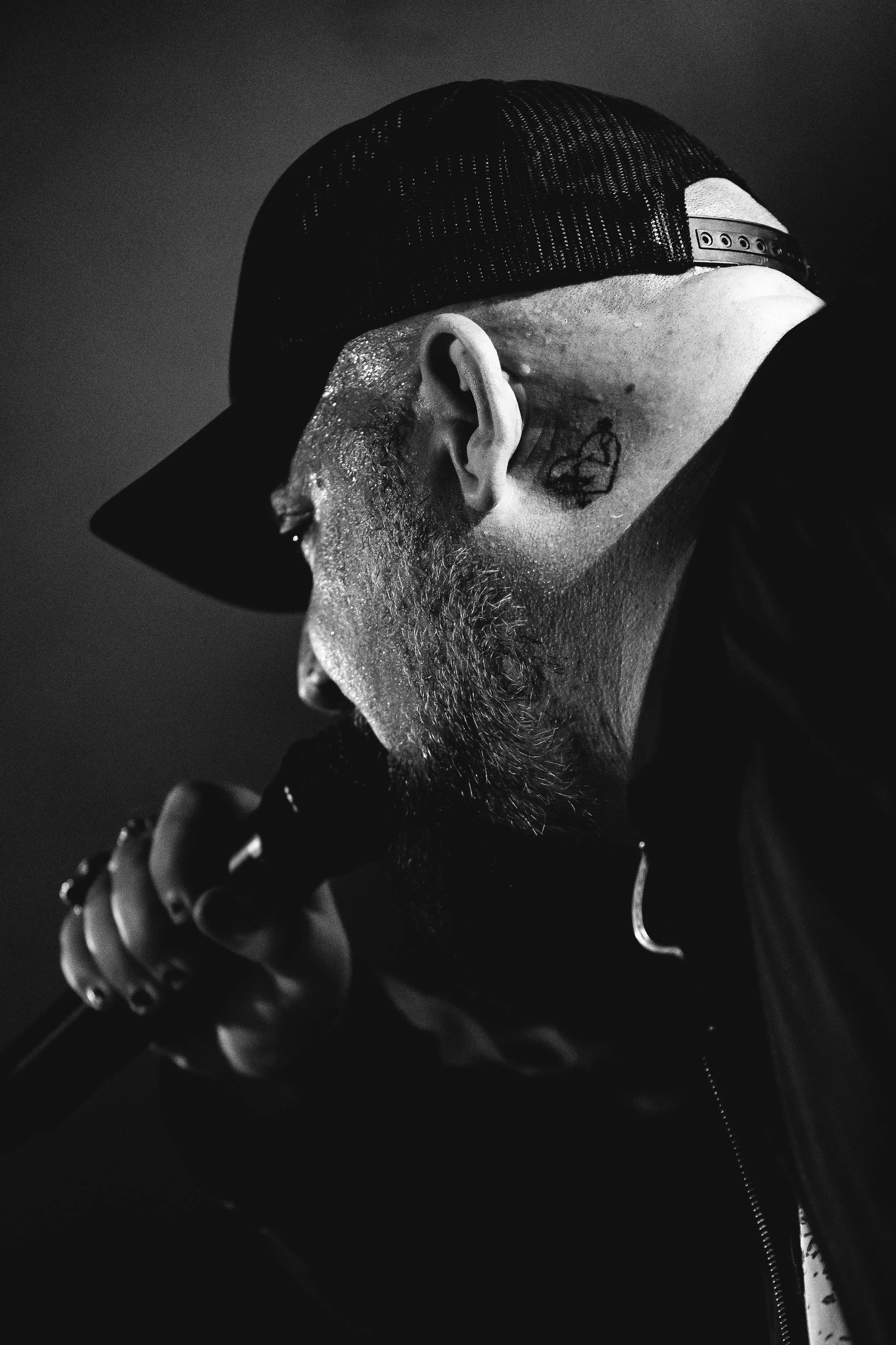 Close-up of a performer (Atreyu) singing into a microphone, wearing a cap backwards, with a beard and tattoos visible on the side of their head, in black and white.