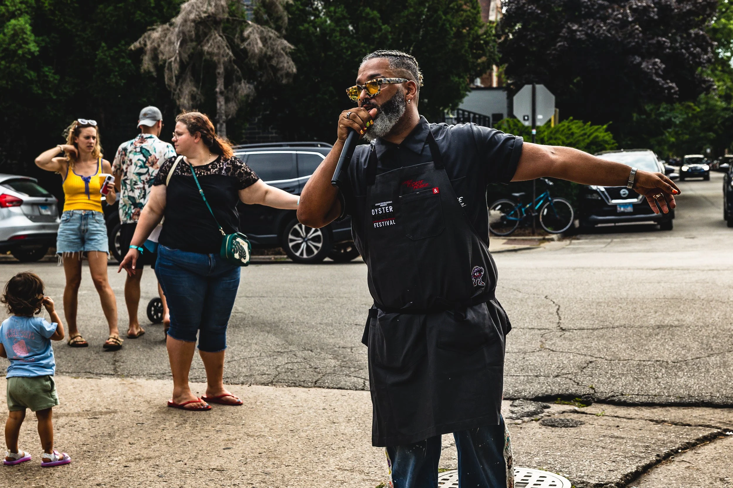 A man with gray hair and beard, wearing sunglasses, a black apron, and black clothes, is performing as a street performer or singer on a sidewalk, holding a microphone. Several people, including women and children, are standing nearby, some looking a
