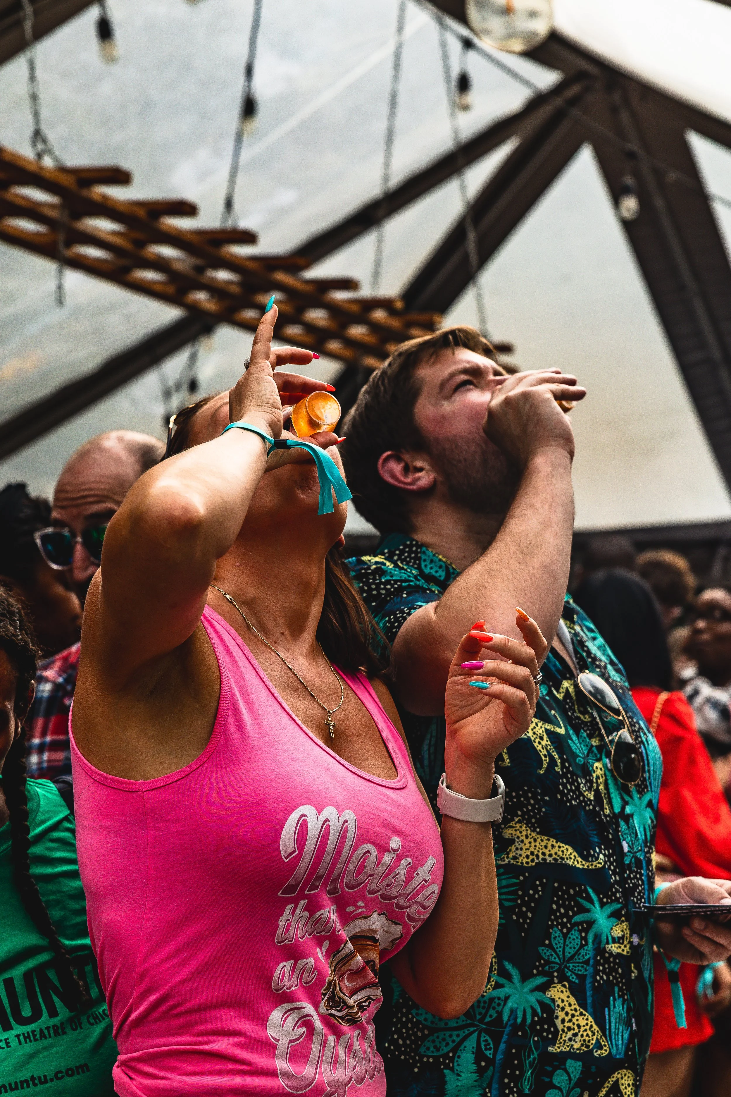 People at a party or concert, some with drinks, standing under a tent or canopy, enjoying the event.