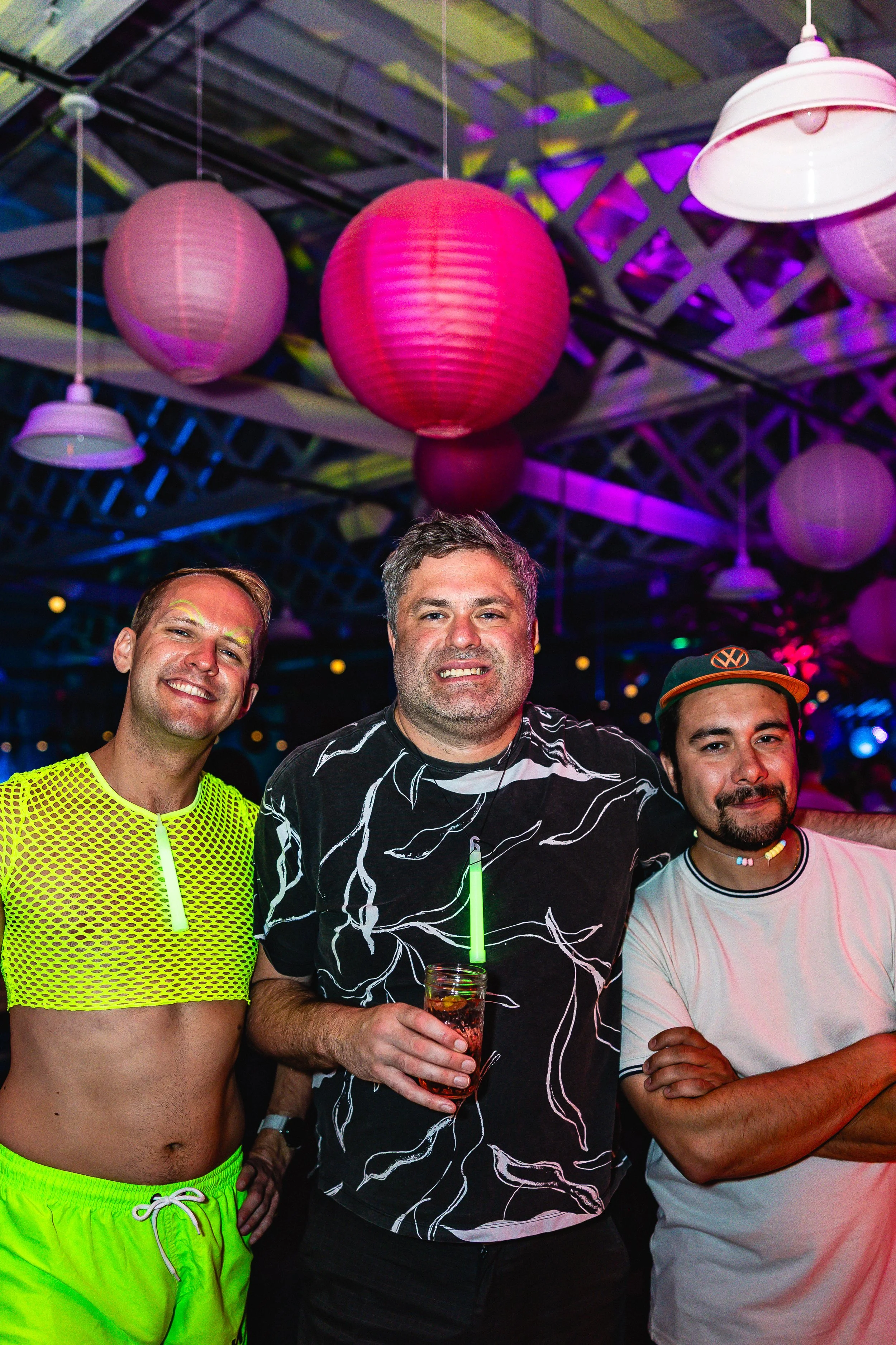 Three men standing close together at a party under colorful hanging lanterns, smiling and posing for the camera.