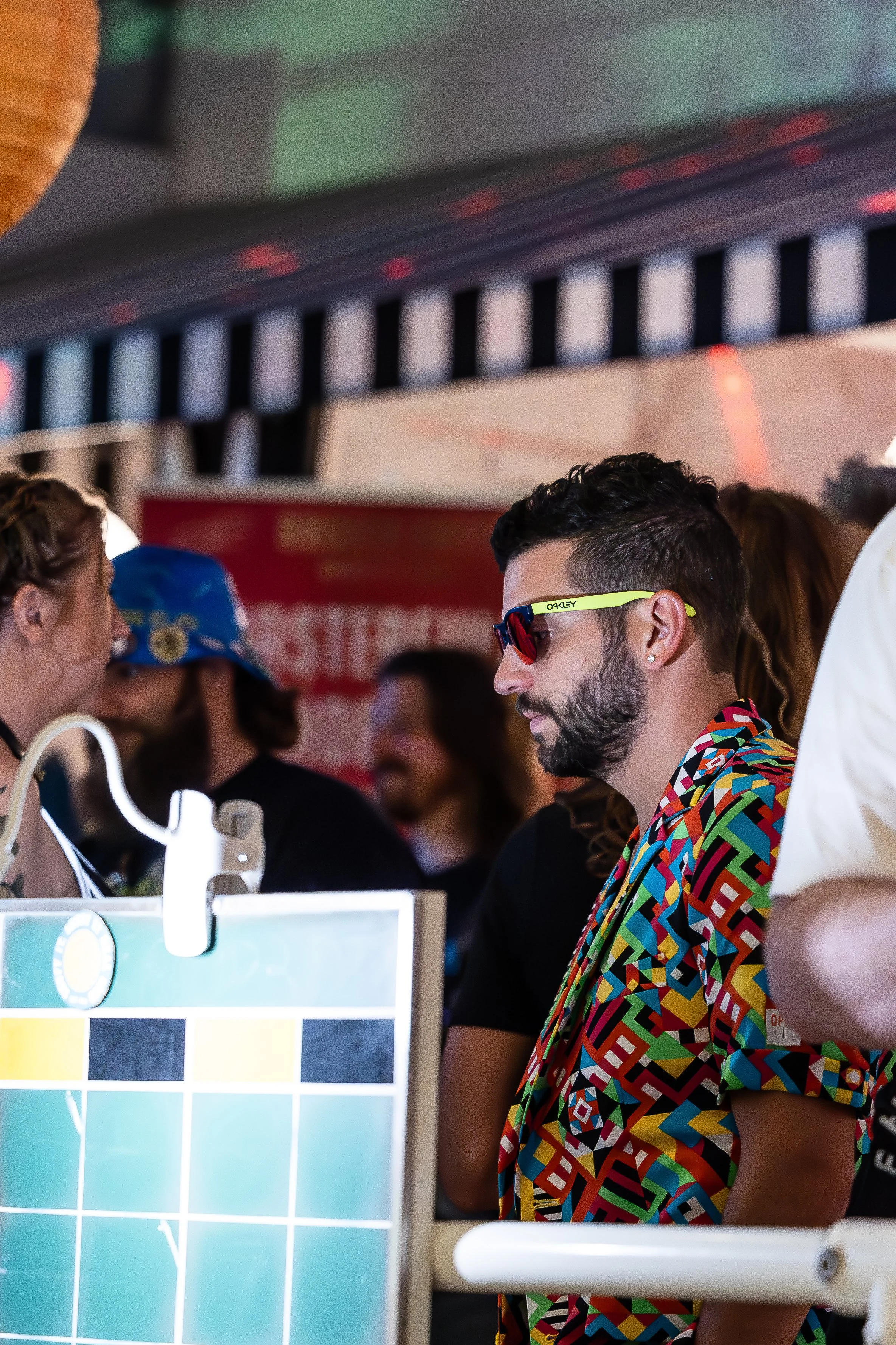 A man with dark hair and a beard wearing colorful, geometric-patterned shirt and red Oakley sunglasses at a crowded event or festival.