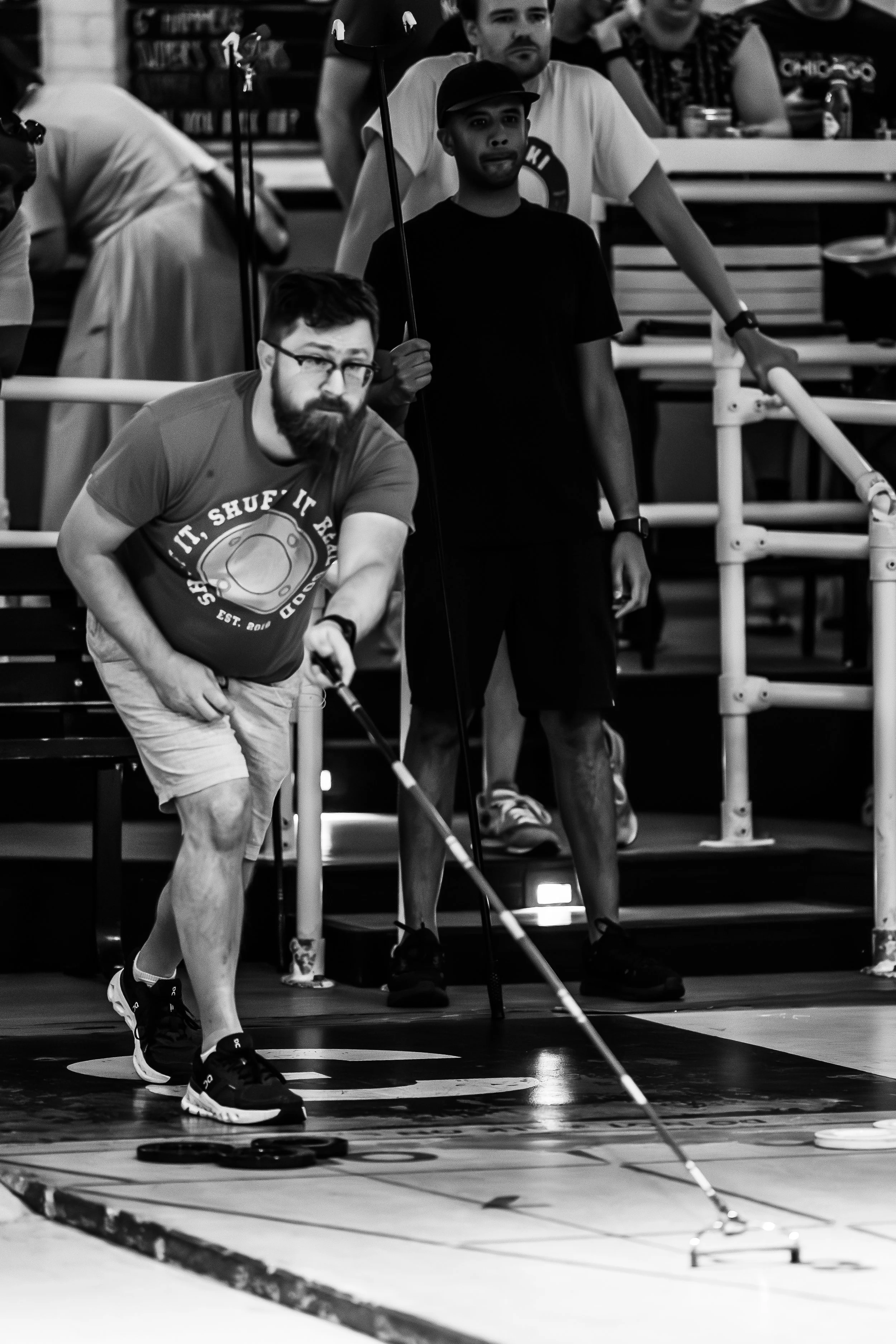 A man with glasses, a beard, and a t-shirt with the text "SHUFF IT" is playing floor shuffleboard, using a stick to slide pucks on the court. Another man in a black t-shirt, shorts, and a cap stands behind him, watching. Several people sit on bleache