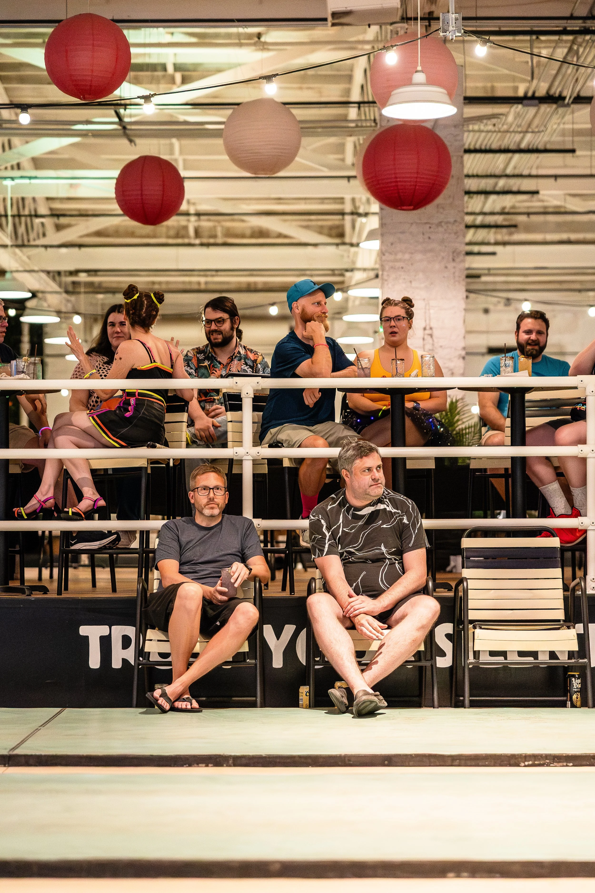 Group of people sitting on a raised platform at an indoor event, some looking at the stage and others conversing, with hanging paper lanterns and string lights overhead.
