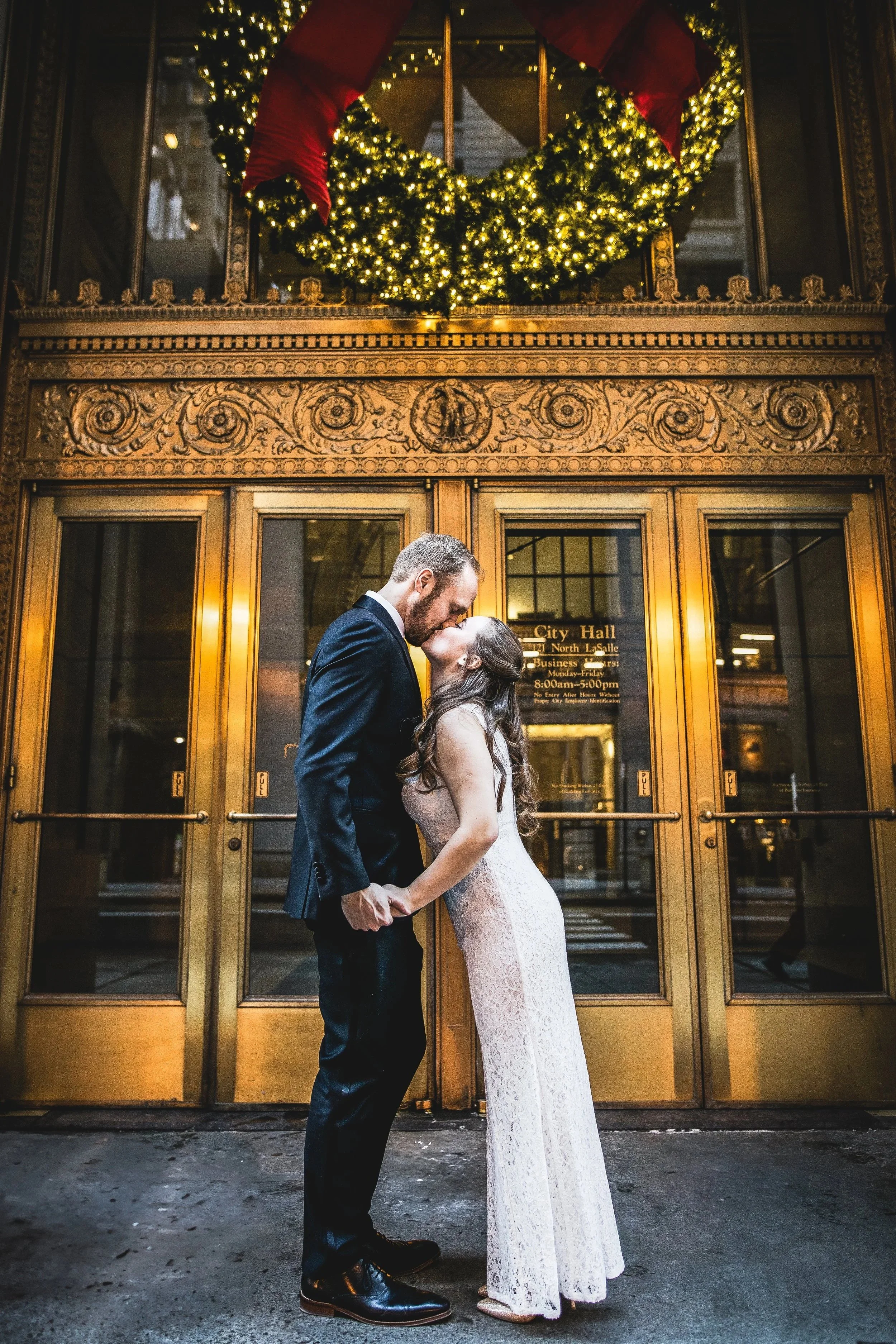 A couple dressed in formal wedding attire sharing a kiss in front of the entrance to a building, with festive Christmas wreath and lights above.