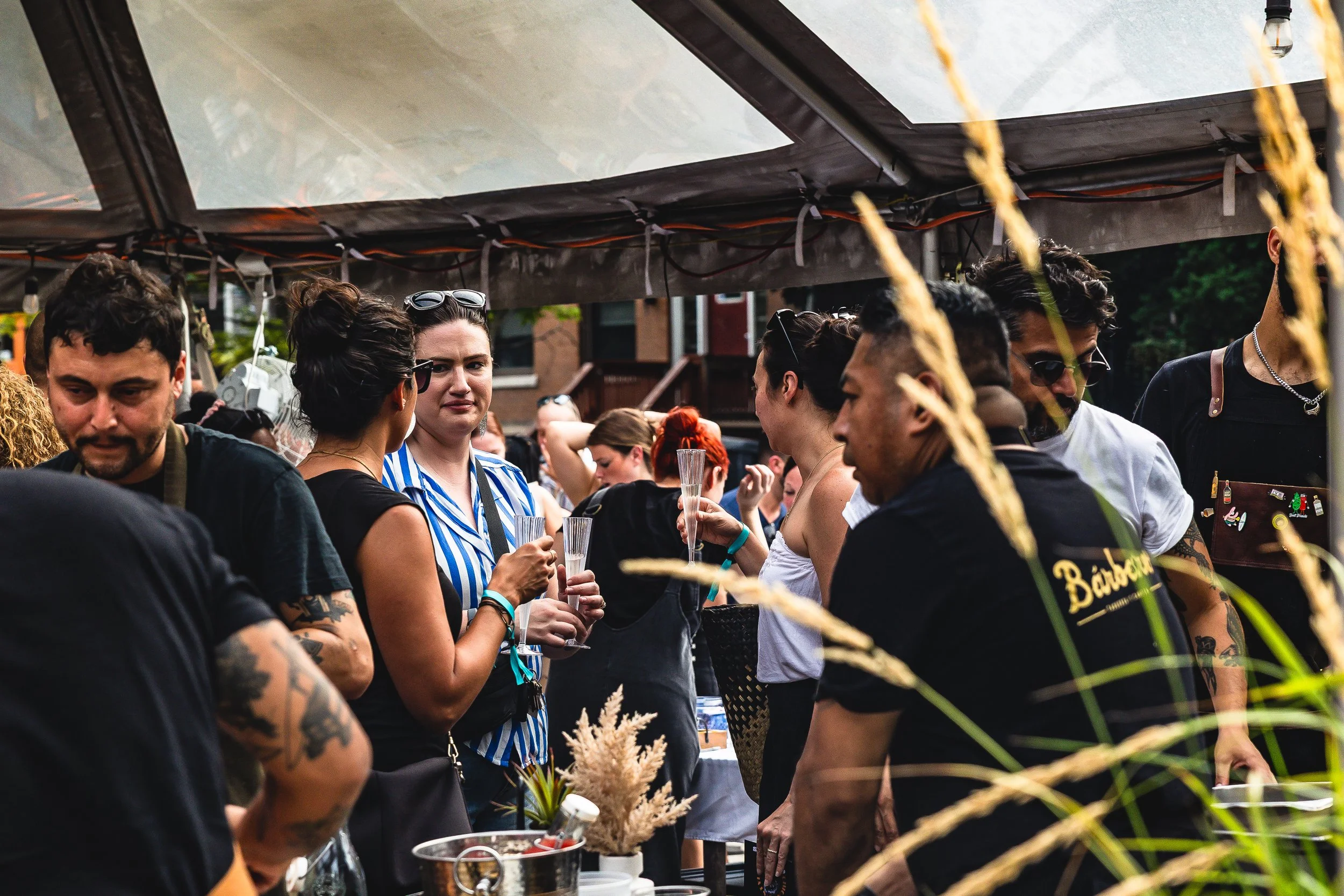 Group of people at an outdoor gathering, some holding glasses, under a canopy, with buildings in the background.