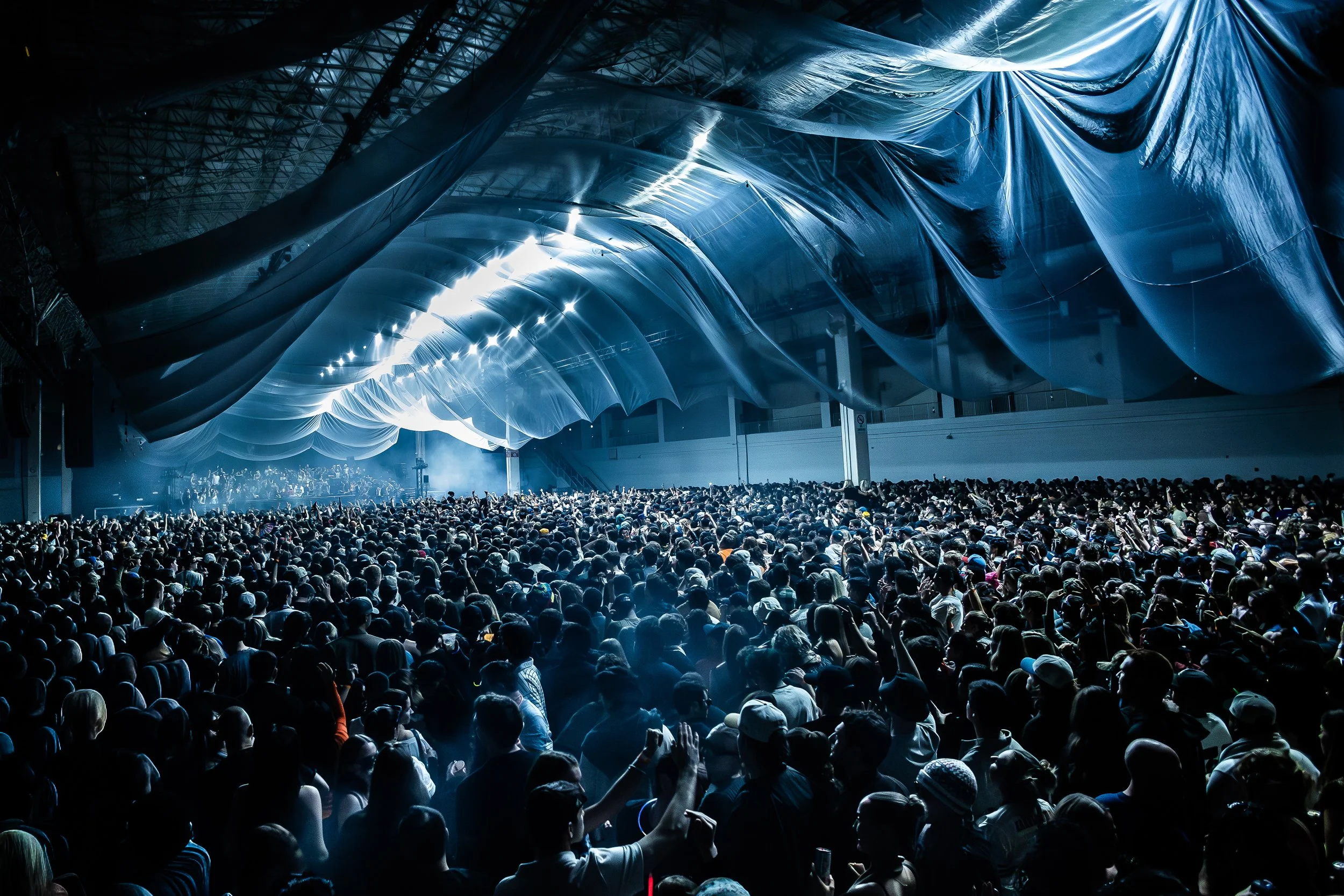 Large indoor concert with a crowd of people facing a stage in a spacious venue with reflective ceiling panels.