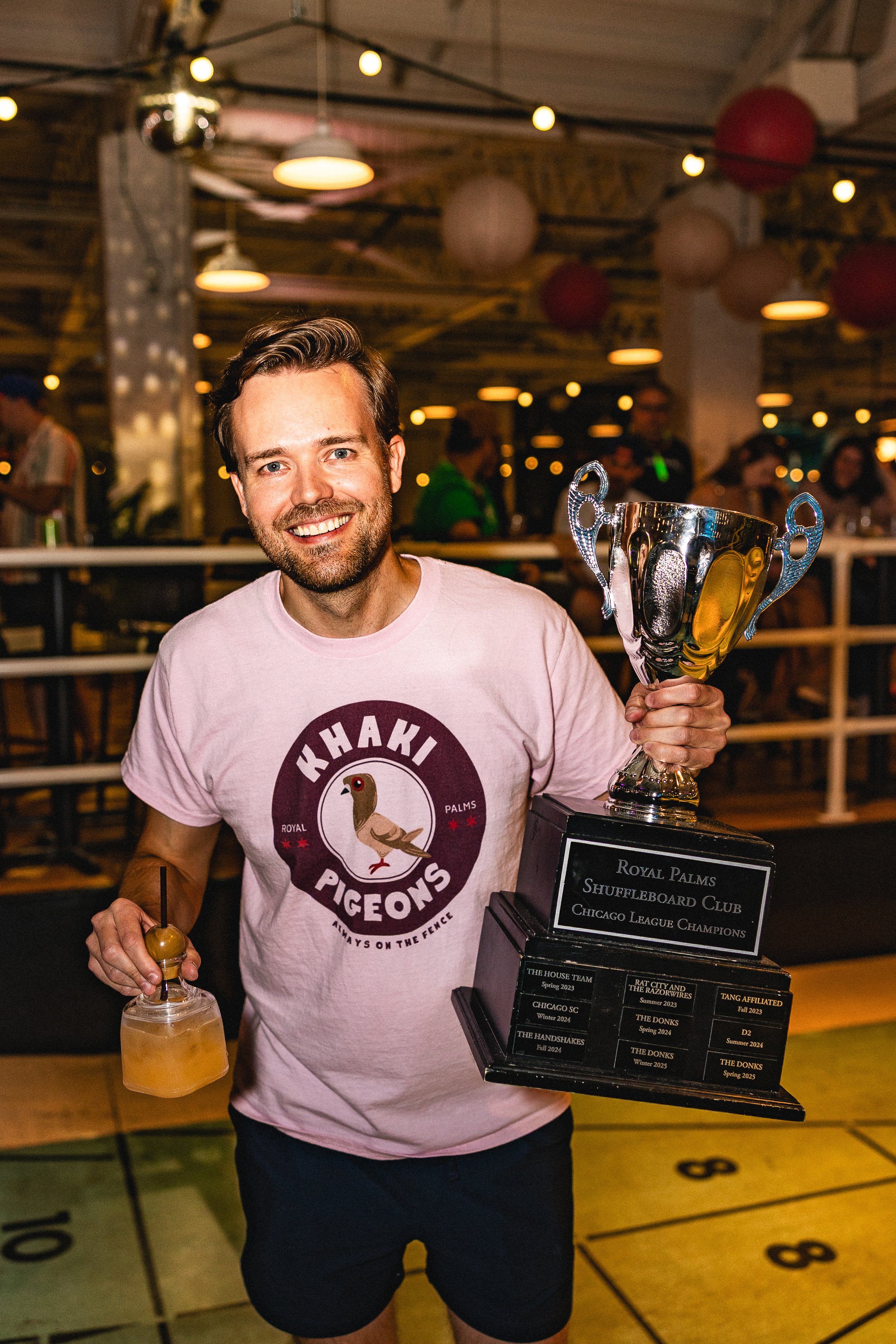 Man smiling and holding a trophy and a drink in a lively indoor venue decorated with paper lanterns, wearing a pink t-shirt with a bird and the text "Khaki Pigeons."