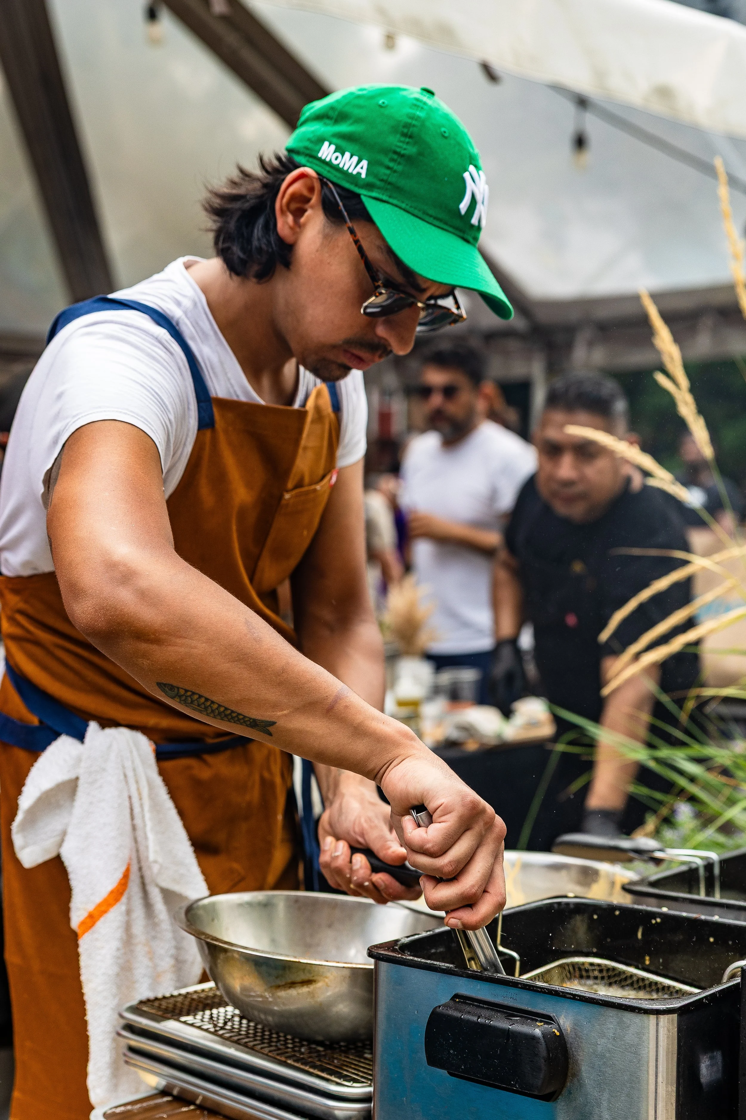 A man wearing a green cap, sunglasses, and a brown apron is frying food with tongs at an outdoor event, while two other men observe in the background.