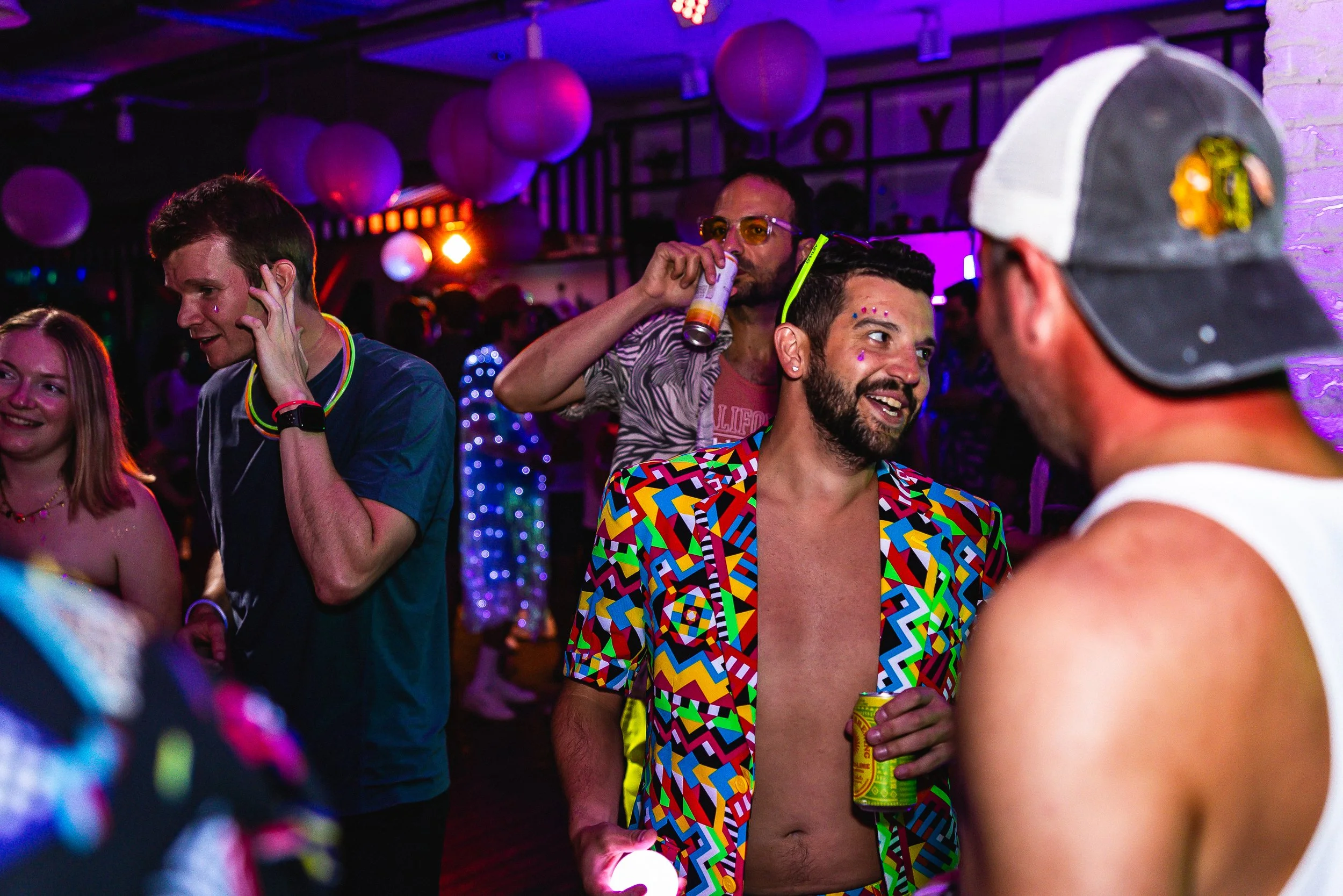 People dancing and socializing at a lively party with colorful lighting. One man is wearing a vibrant, geometric-patterned jacket and holding a can, chatting with another person in a white tank top and a Donald Trump hat. The background includes purp
