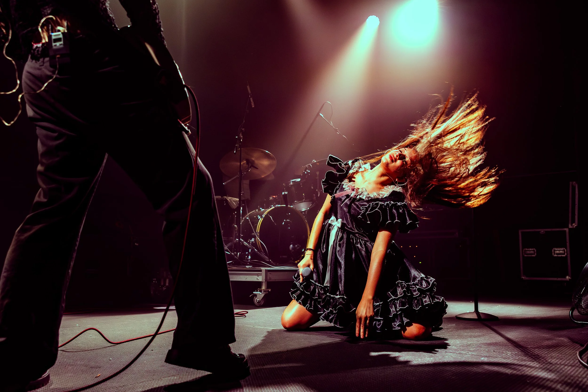A female singer in a ruffled black dress kneels on stage, holding a microphone, with her hair flying while performing passionately. A musician on the left is partly visible, and drums are seen in the background with stage lights shining down.