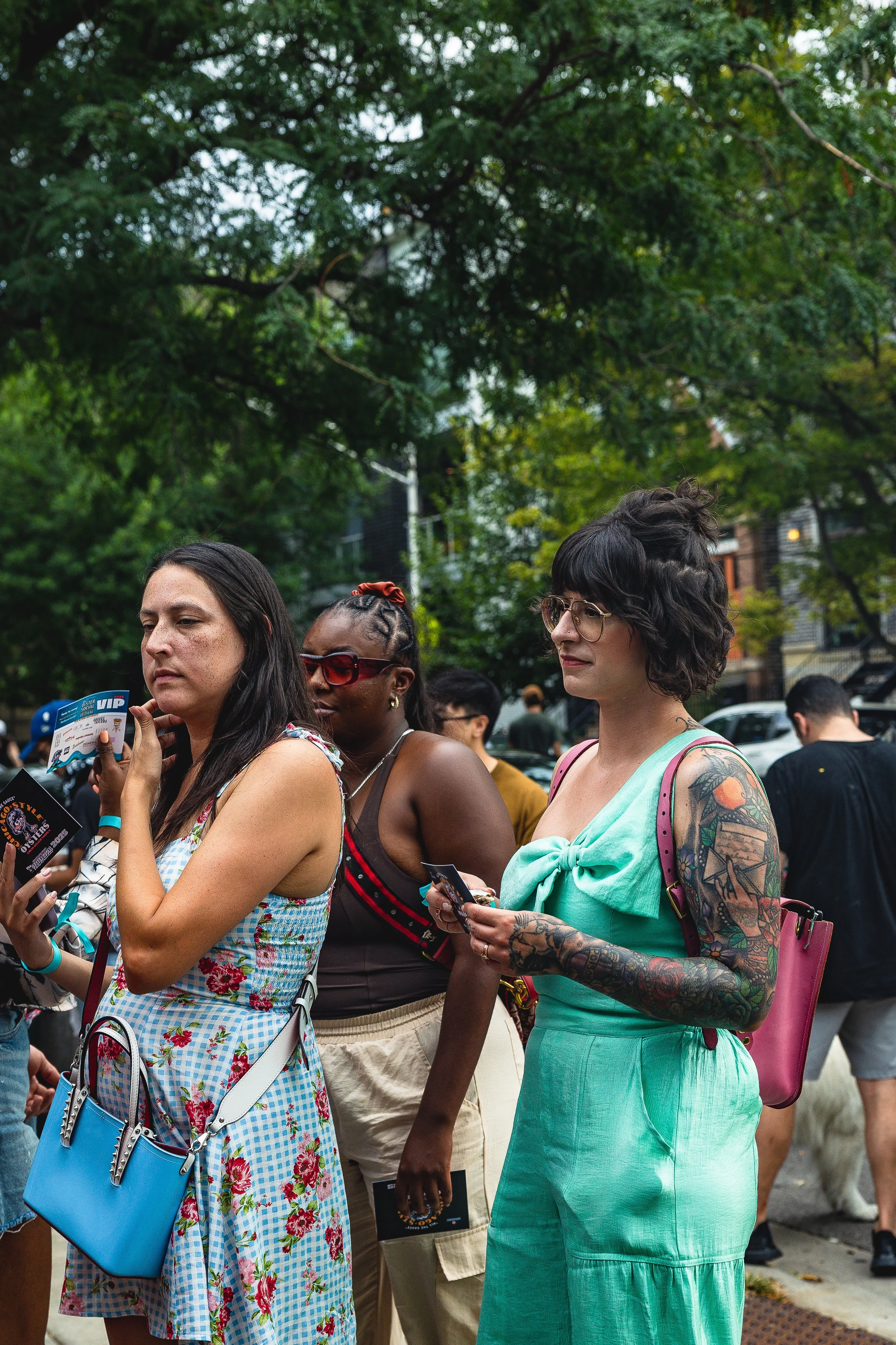 Three women standing in a crowd outdoors, each holding a ticket or pamphlet, with trees and other people in the background.