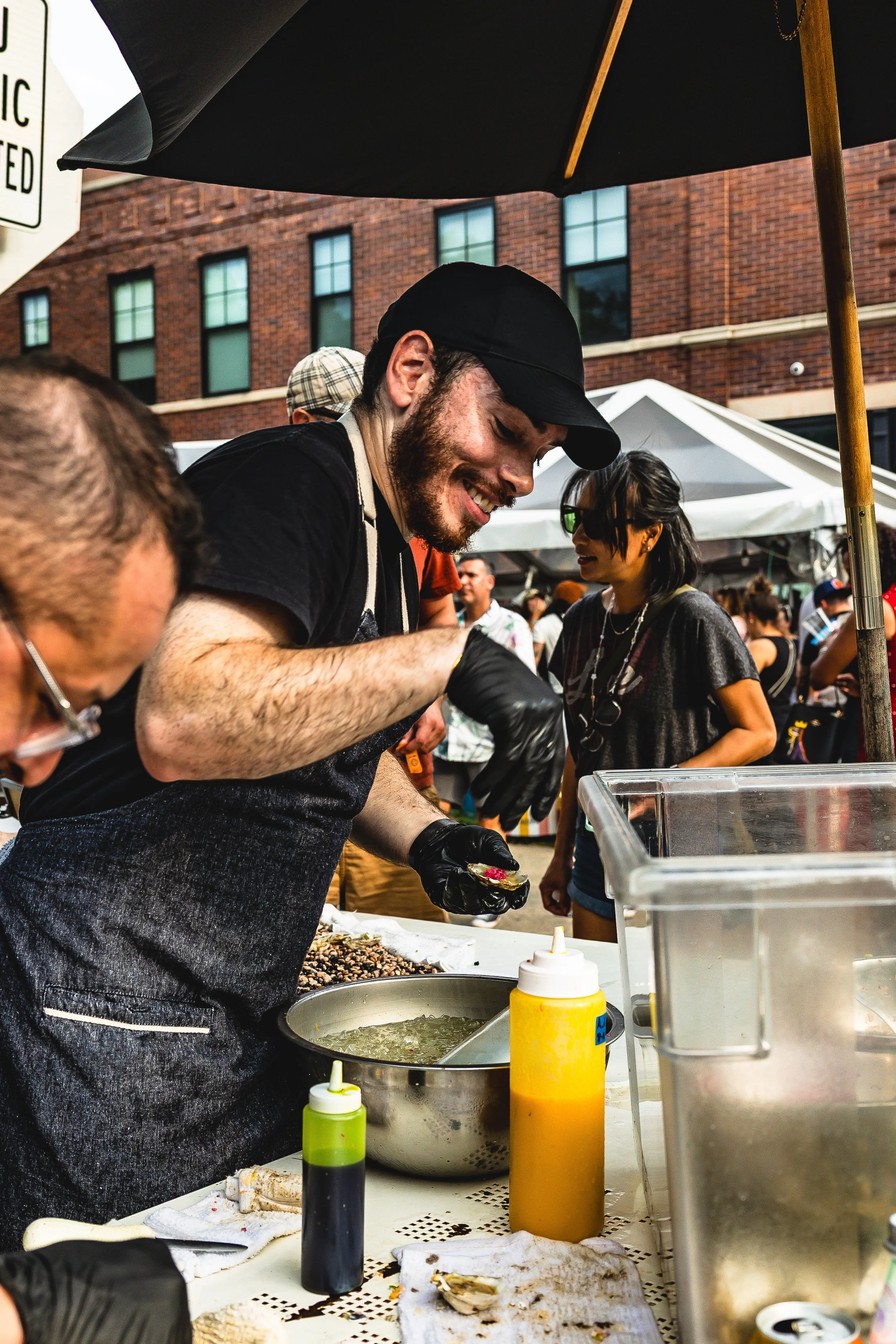 A man wearing a black cap, black gloves, and a black apron is smiling while preparing food at a street food stall at an outdoor market, with people and tents in the background.