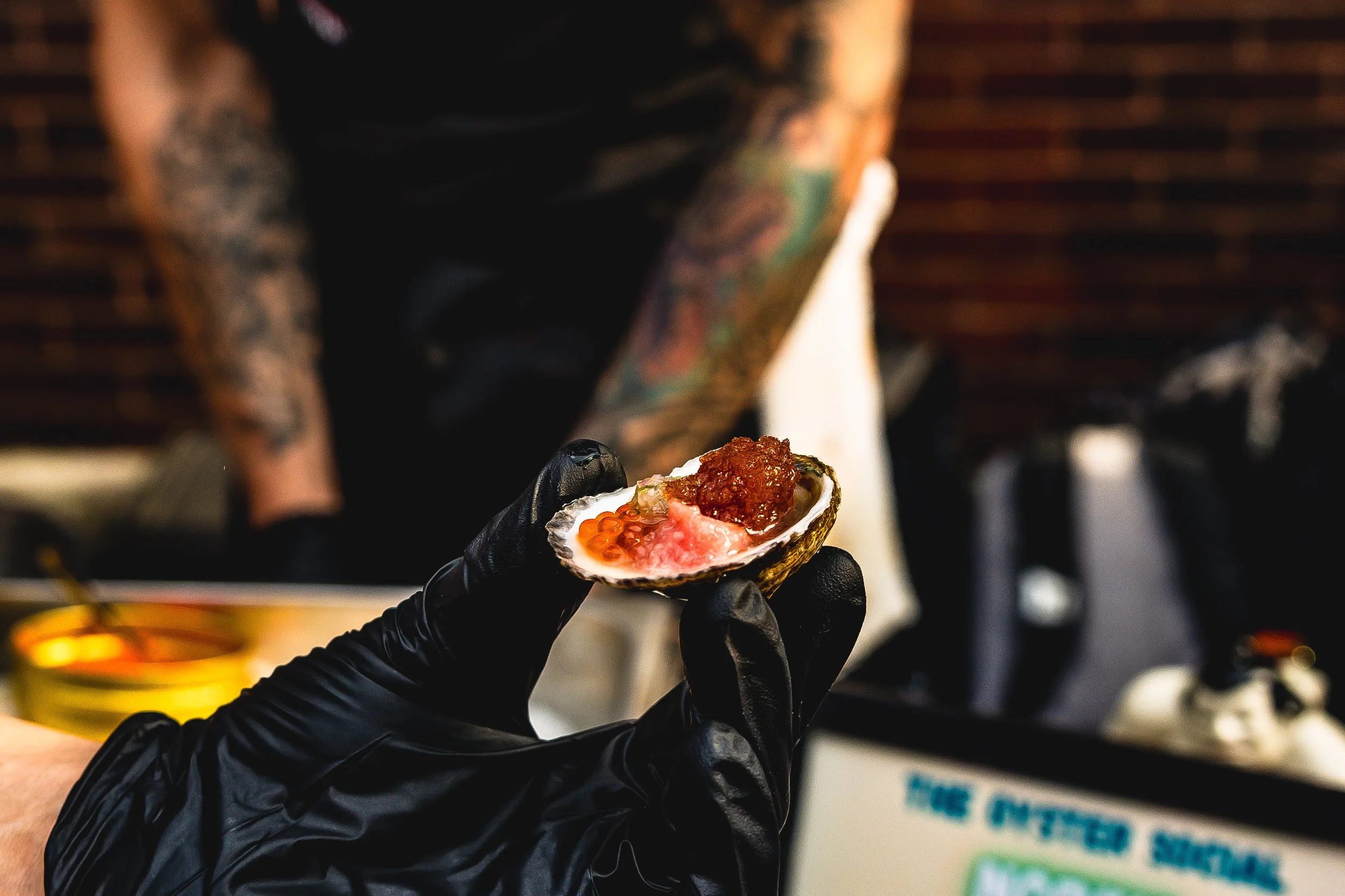 A gloved hand holding a fresh oyster shell filled with orange fish roe, pink shrimp, and brownish topping, with a tattooed person in the background.