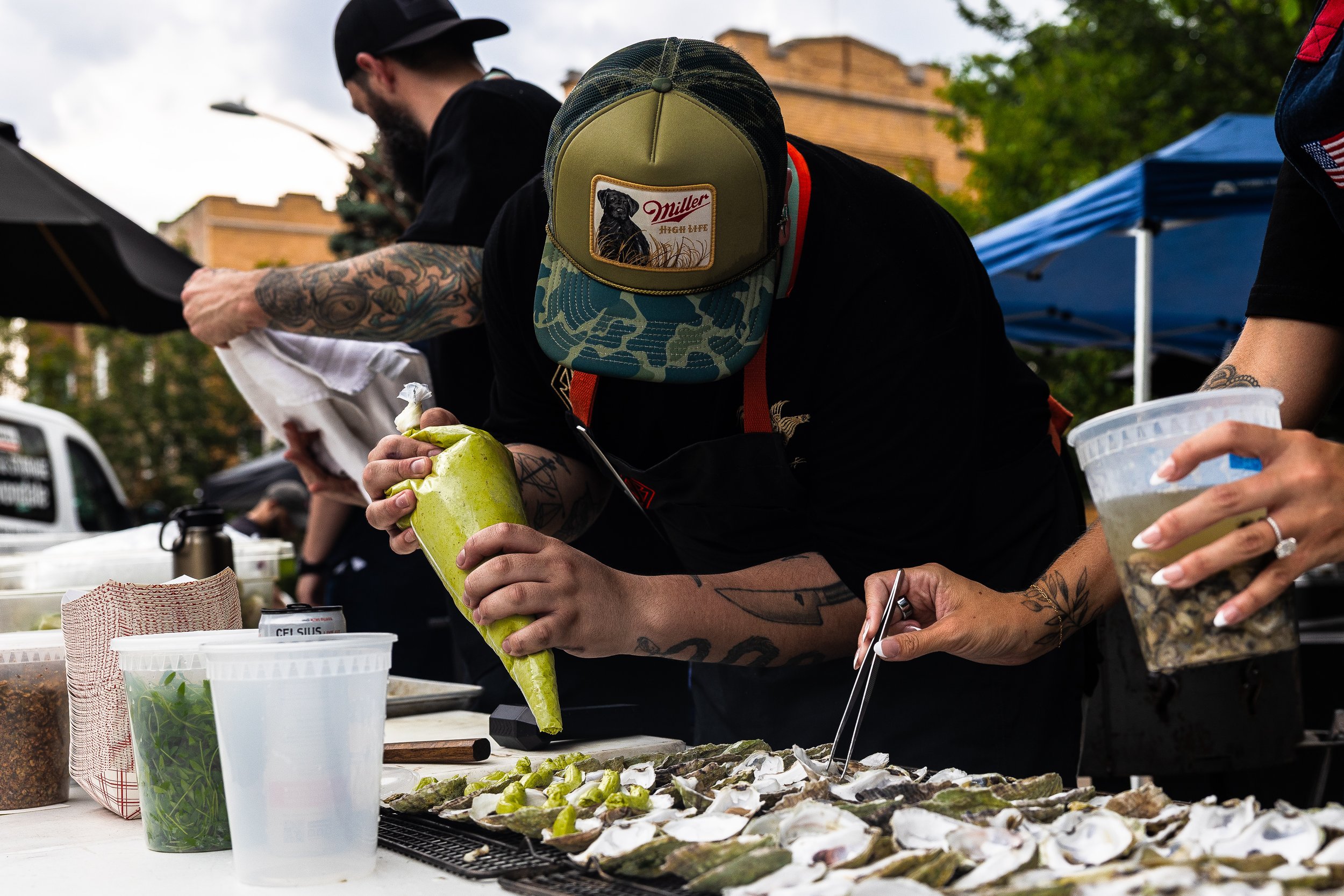 People at an outdoor food stall preparing oysters, with one person squeezing lemon over them, surrounded by containers of condiments and toppings.