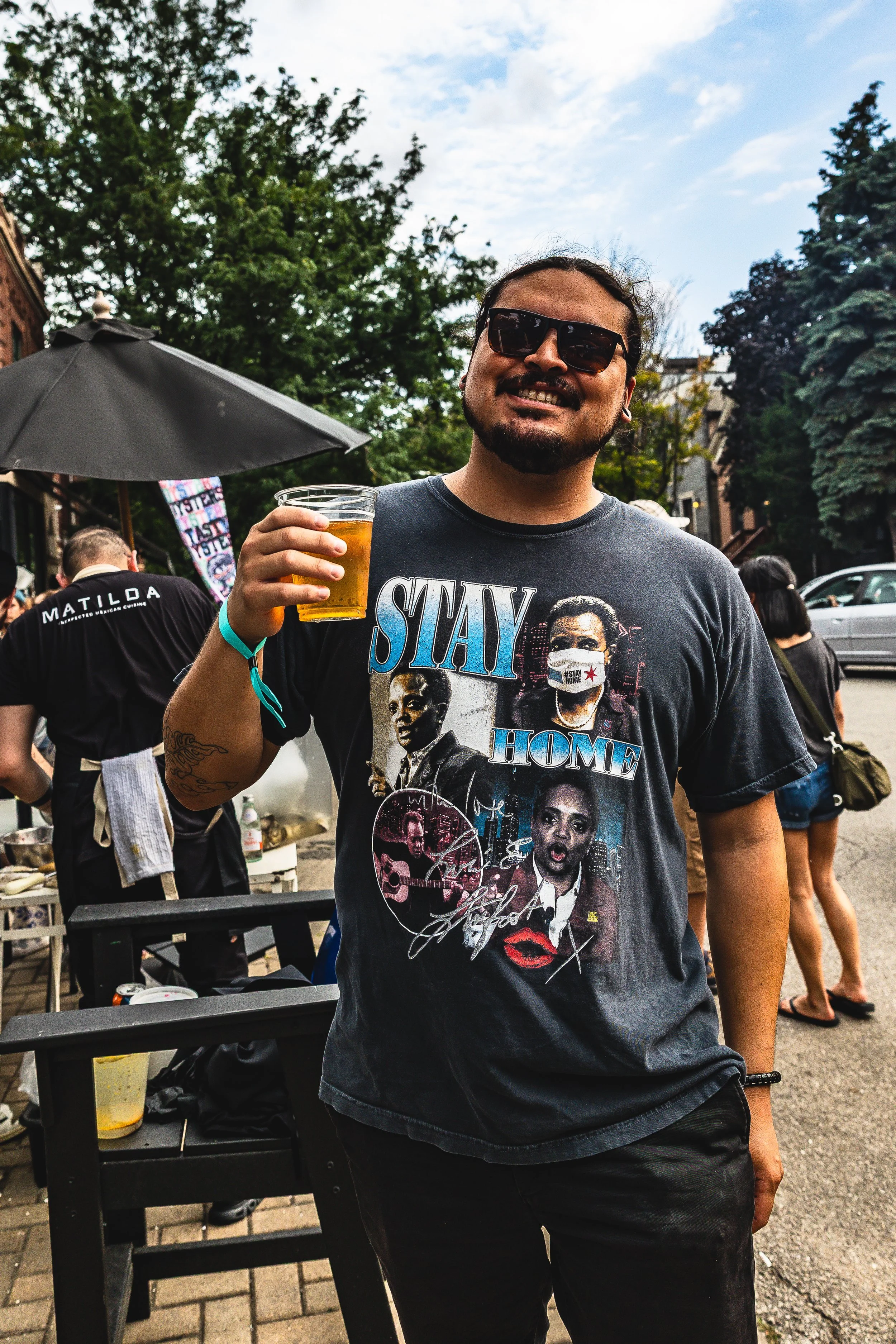 A man wearing dark sunglasses and a black T-shirt with graphics and the words 'Stay Home' is smiling and holding a drink in a clear plastic cup outdoors during a gathering or event.