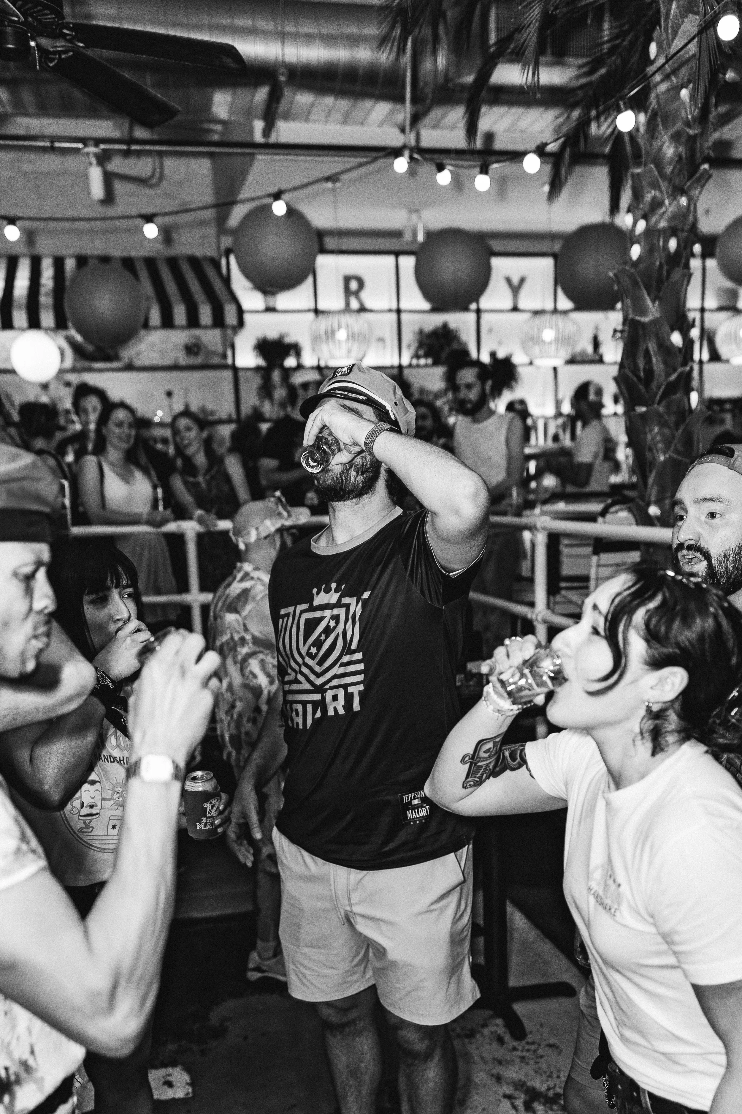 Group of people drinking and socializing at a party with decorative hanging lanterns and string lights, in a lively indoor setting.