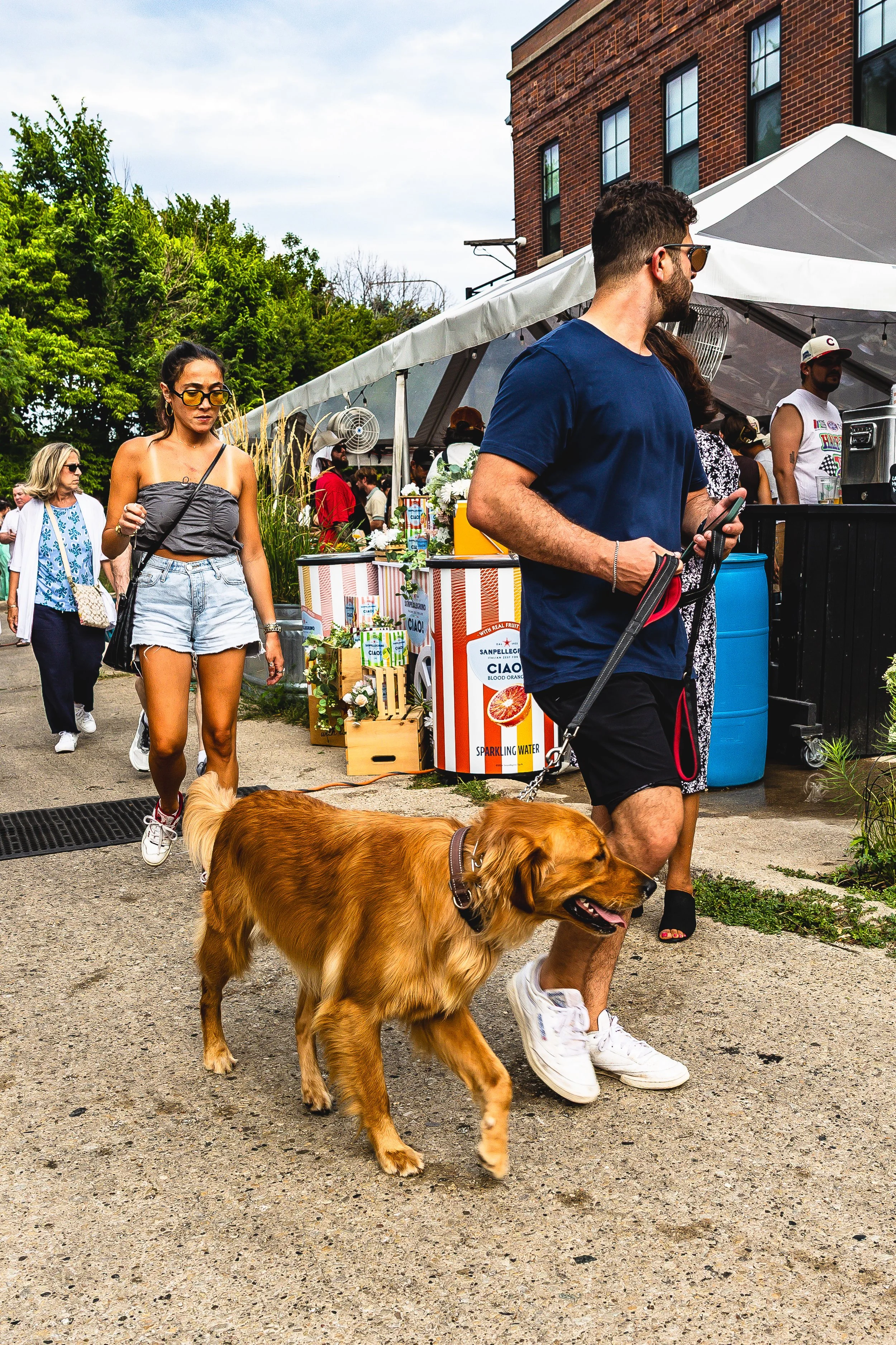 People at an outdoor market or fair, including a man walking a golden retriever dog, a woman in denim shorts and sunglasses, and others near vendor tents with drinks and flowers in city setting.