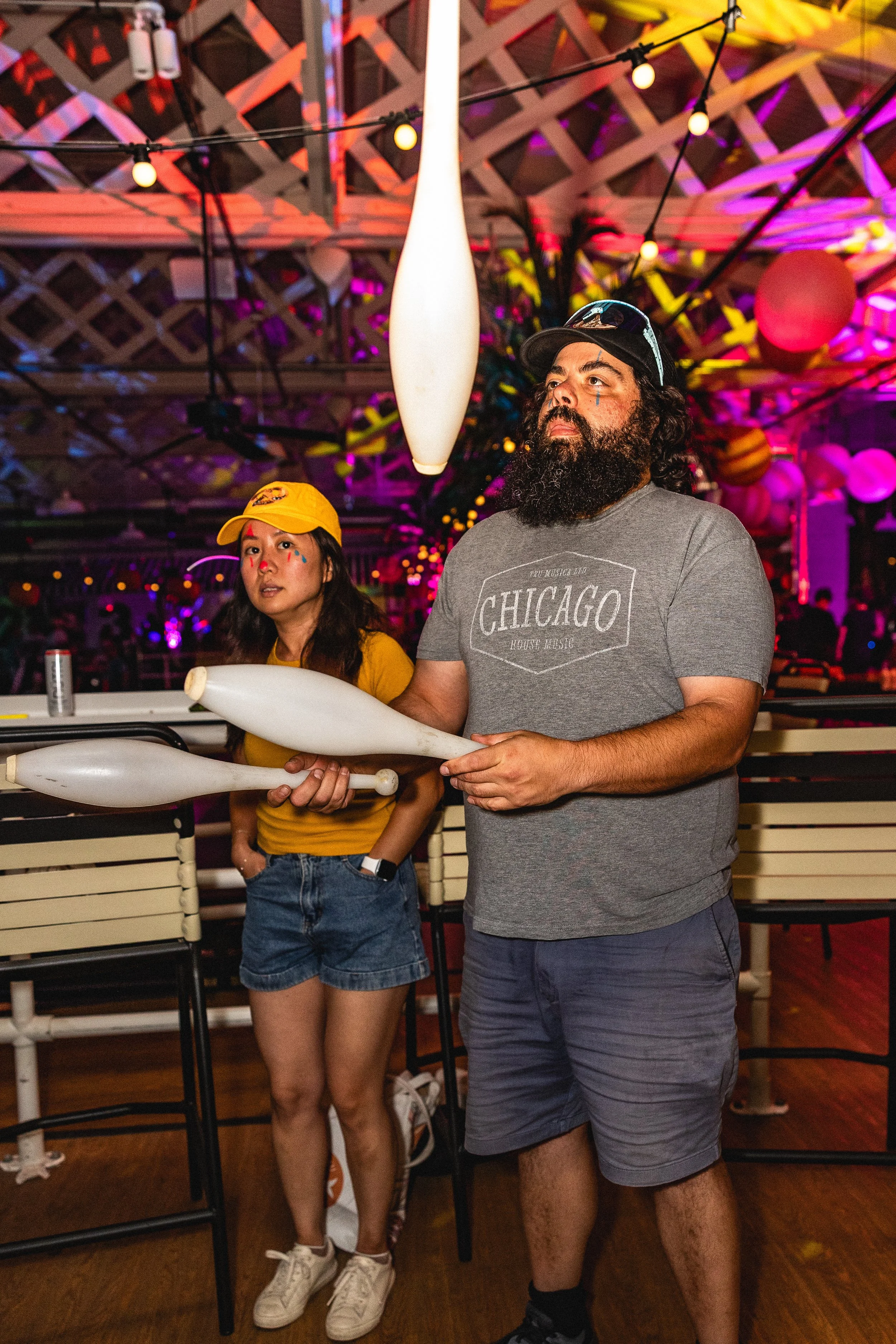 Two people, a woman and a man, standing in a colorful, festive indoor setting with decorative balloons and lighting, the woman is wearing a yellow cap and a yellow shirt, and the man is wearing a gray T-shirt with 'Chicago' written on it and a black 