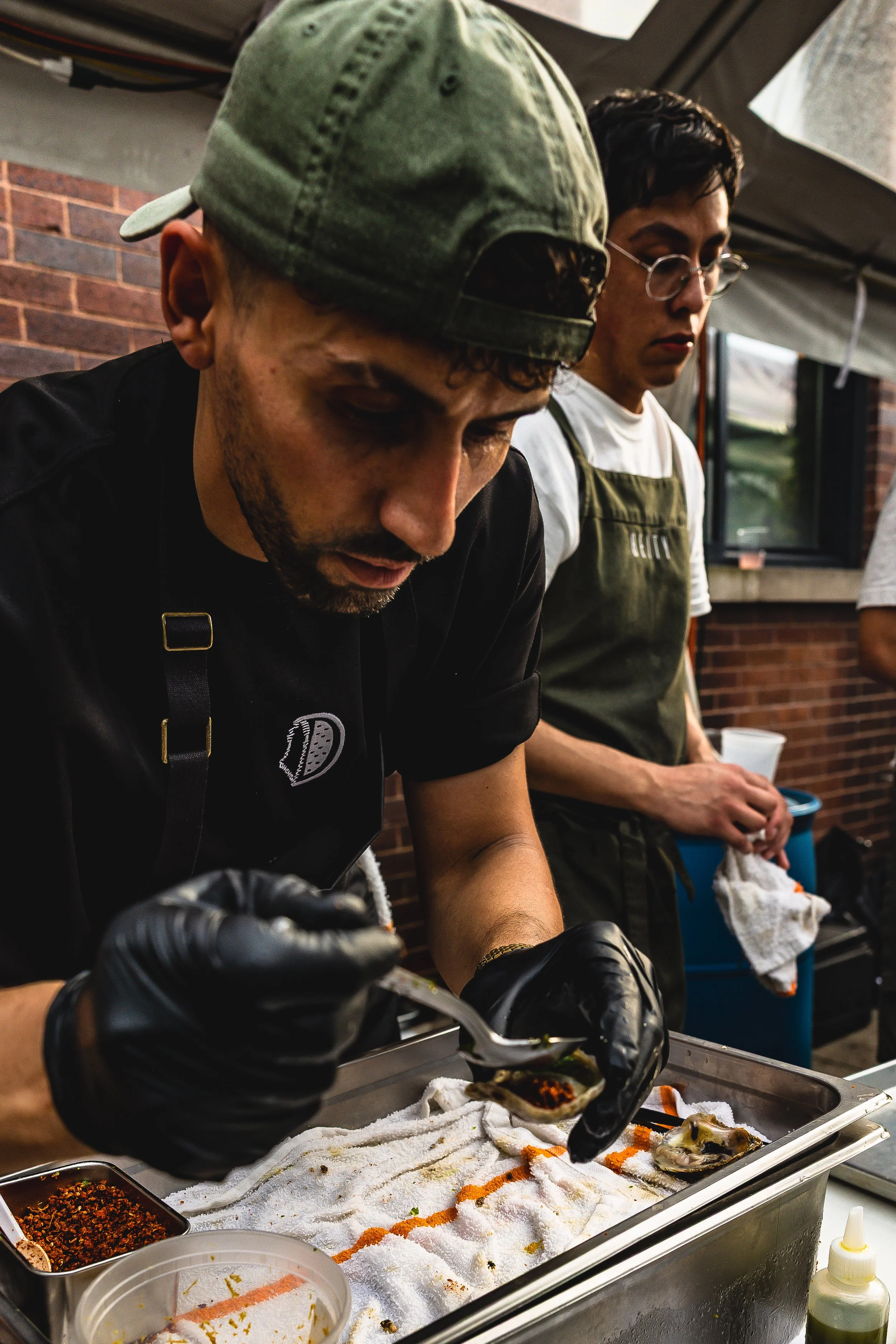 Two people preparing food in an outdoor kitchen, with one person wearing a baseball cap and black gloves, focusing on handling an oyster. The other person, in glasses and an apron, stands behind observing, at a brick wall backdrop.