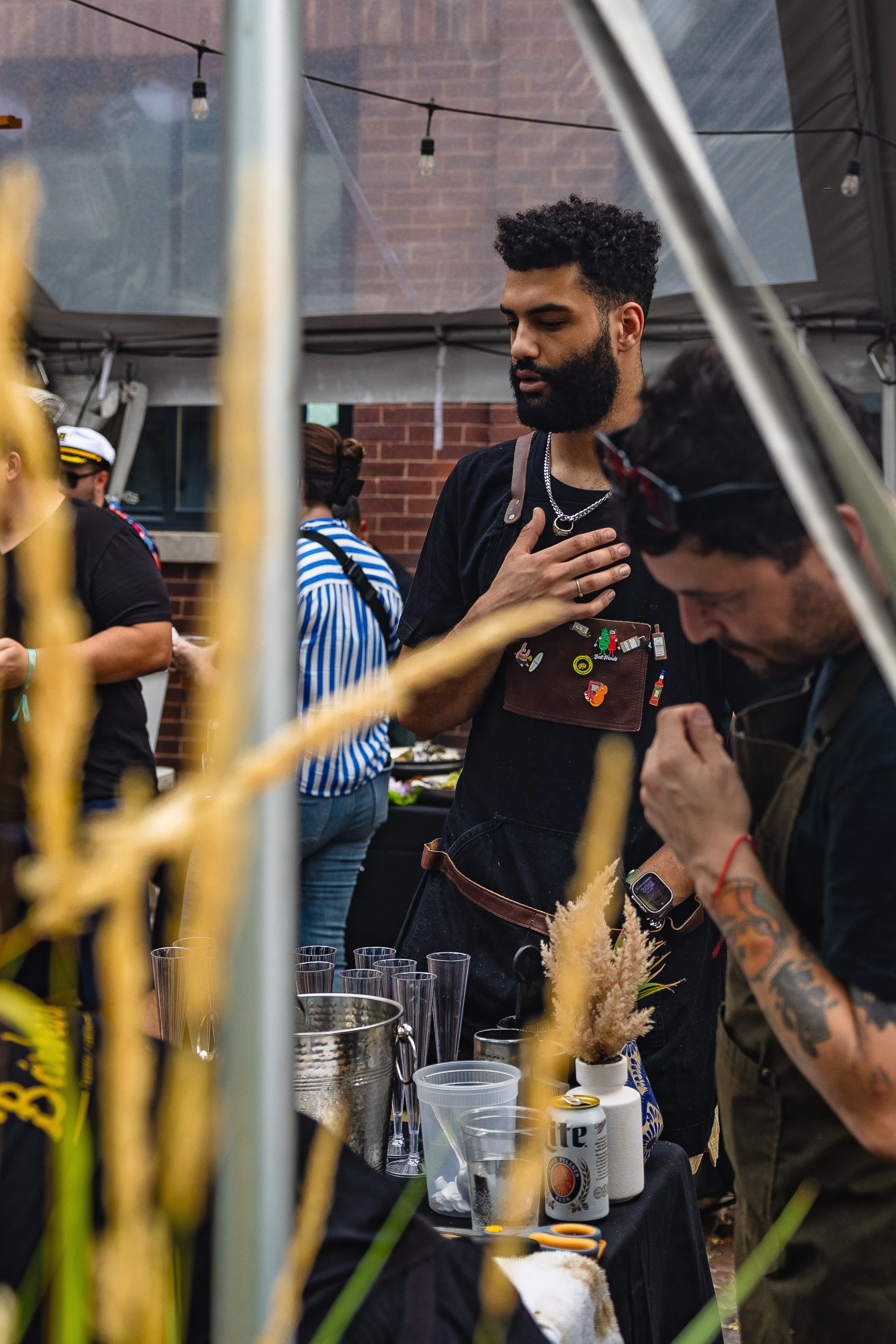 Two men preparing drinks at an outdoor event with tents, string lights, and people in the background.