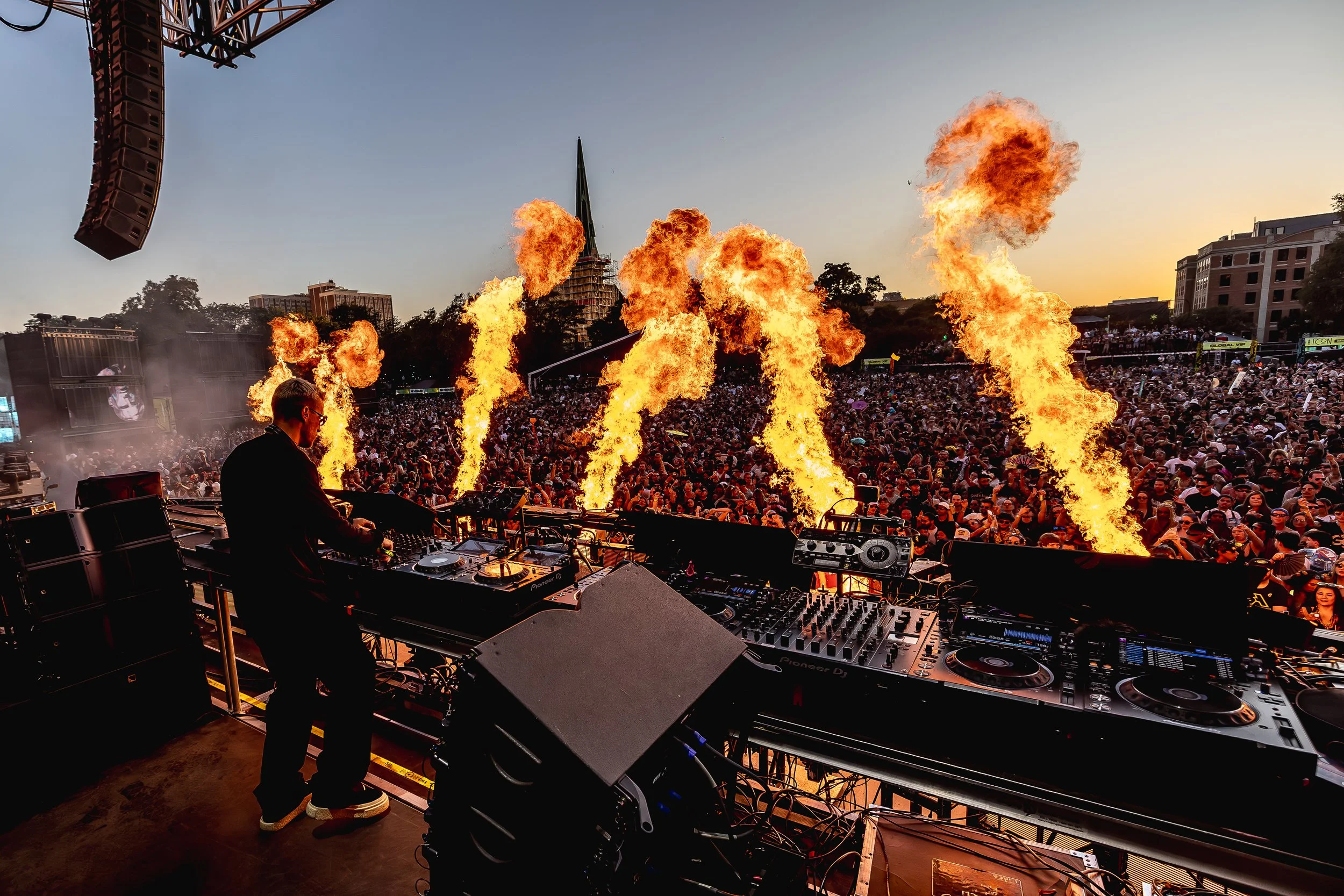 DJ performing on stage with fire effects, large crowd at an outdoor music festival during sunset, city buildings in the background.