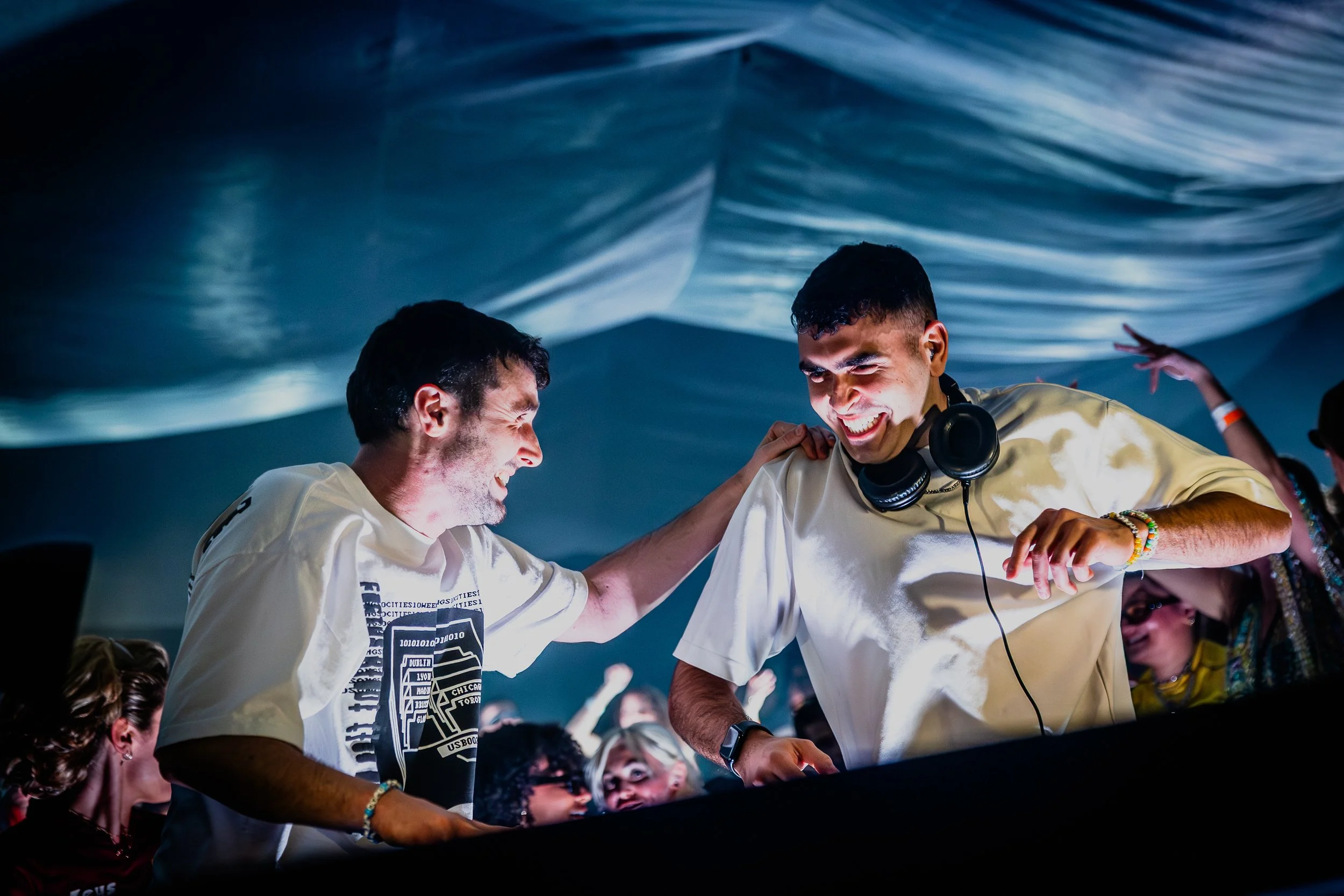 Fred Again and Sammy Vigil smiling and dancing at a party or music event under a large, blue, wavy fabric ceiling. They are enjoying music, with one wearing headphones around his neck. The crowd is visible in the background.