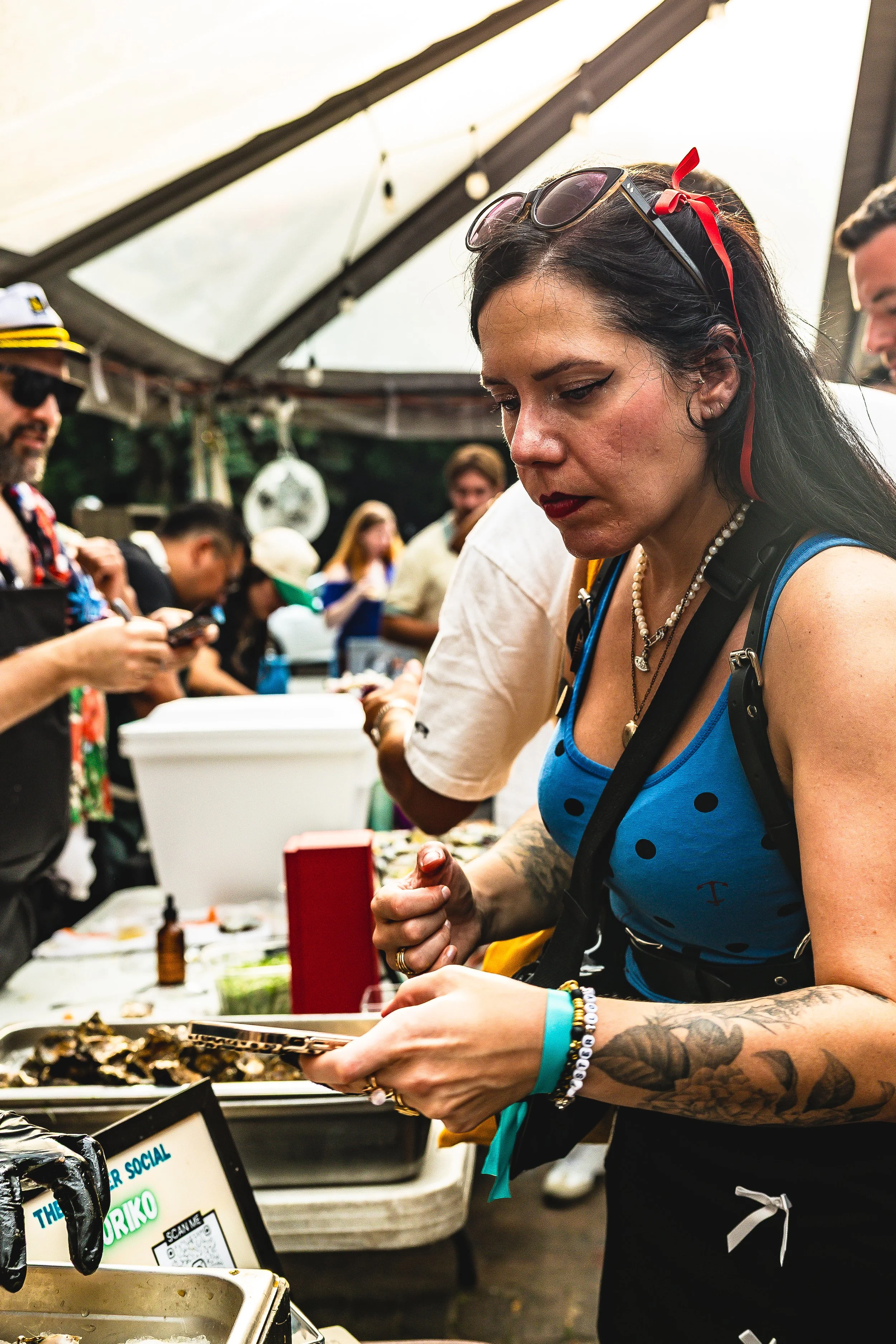 Woman with black hair, sunglasses, and tattoos, wearing a blue top, at an outdoor event, looking at a tray of food on a table.