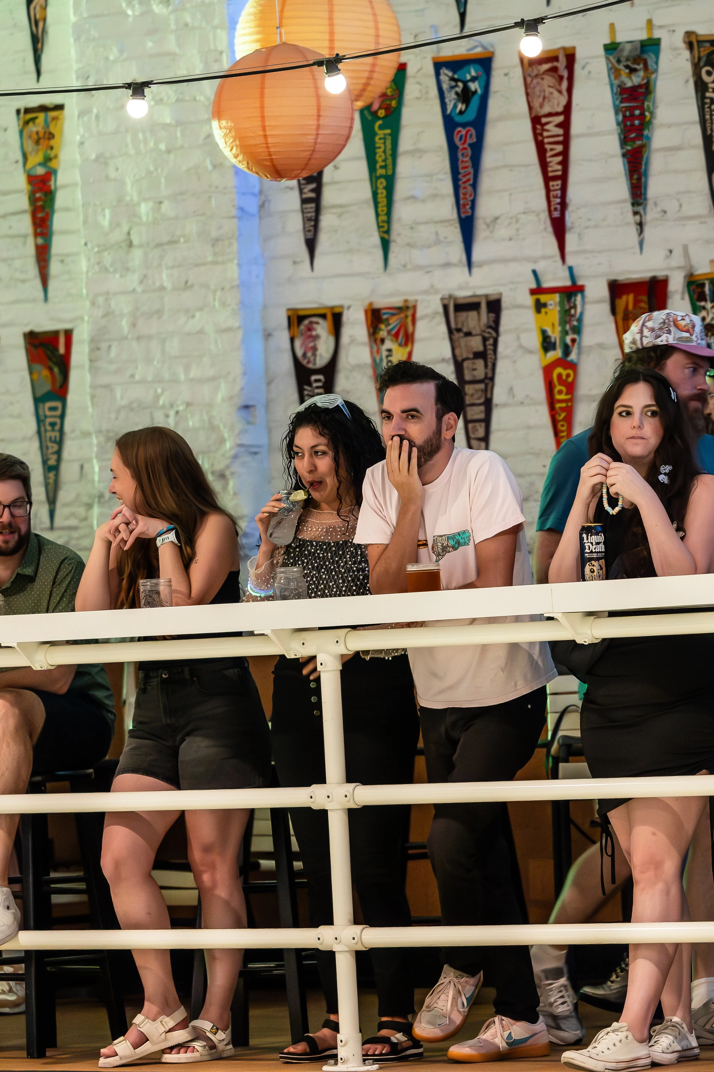 Group of people standing at a bar or railing, watching an event, with colorful banners hanging on a white brick wall behind them. Some are smiling, drinking, or resting their heads on their hands, in a casual event setting.