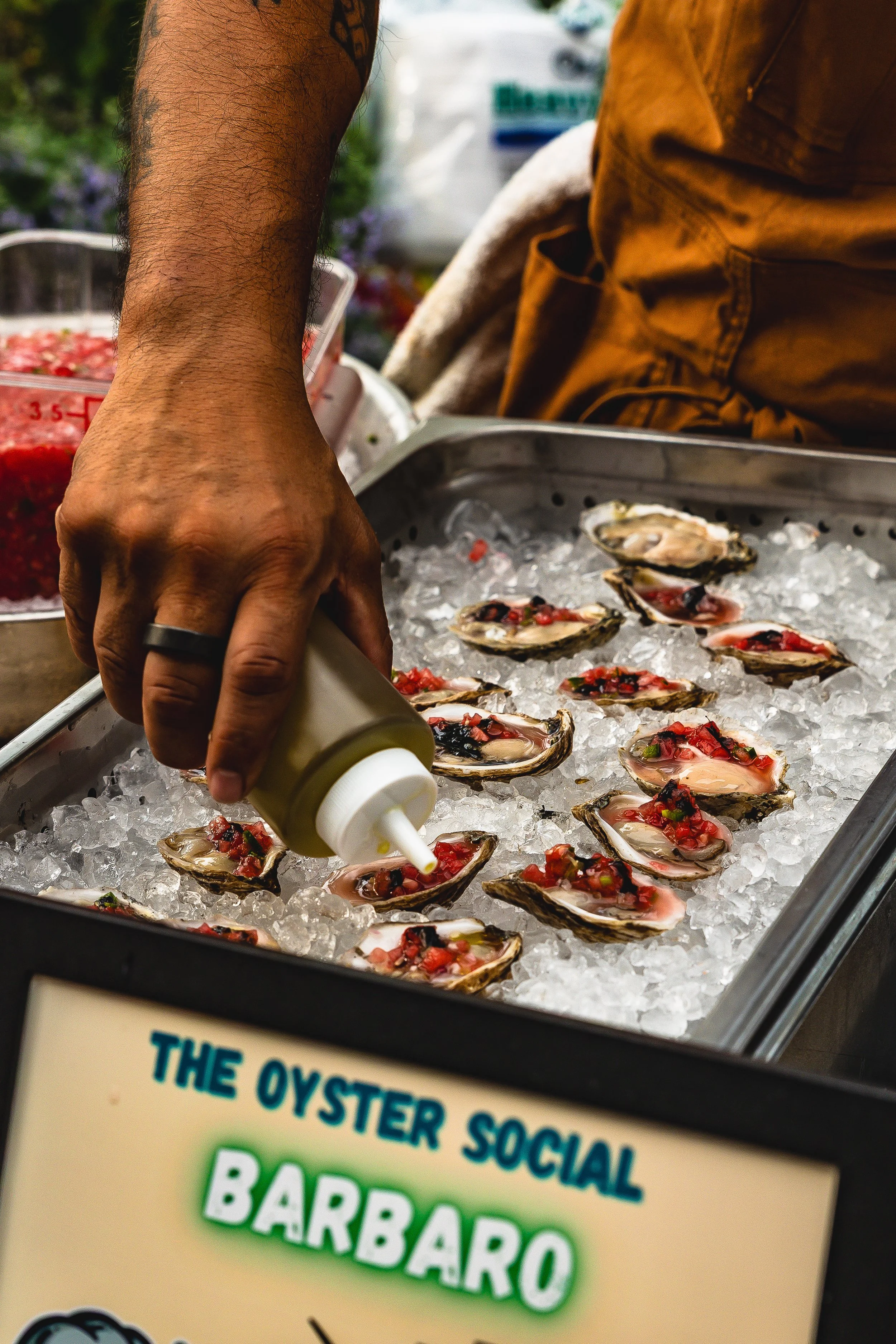 Person drizzling sauce over oysters on ice at a seafood stand labeled 'The Oyster Social Barraro.'
