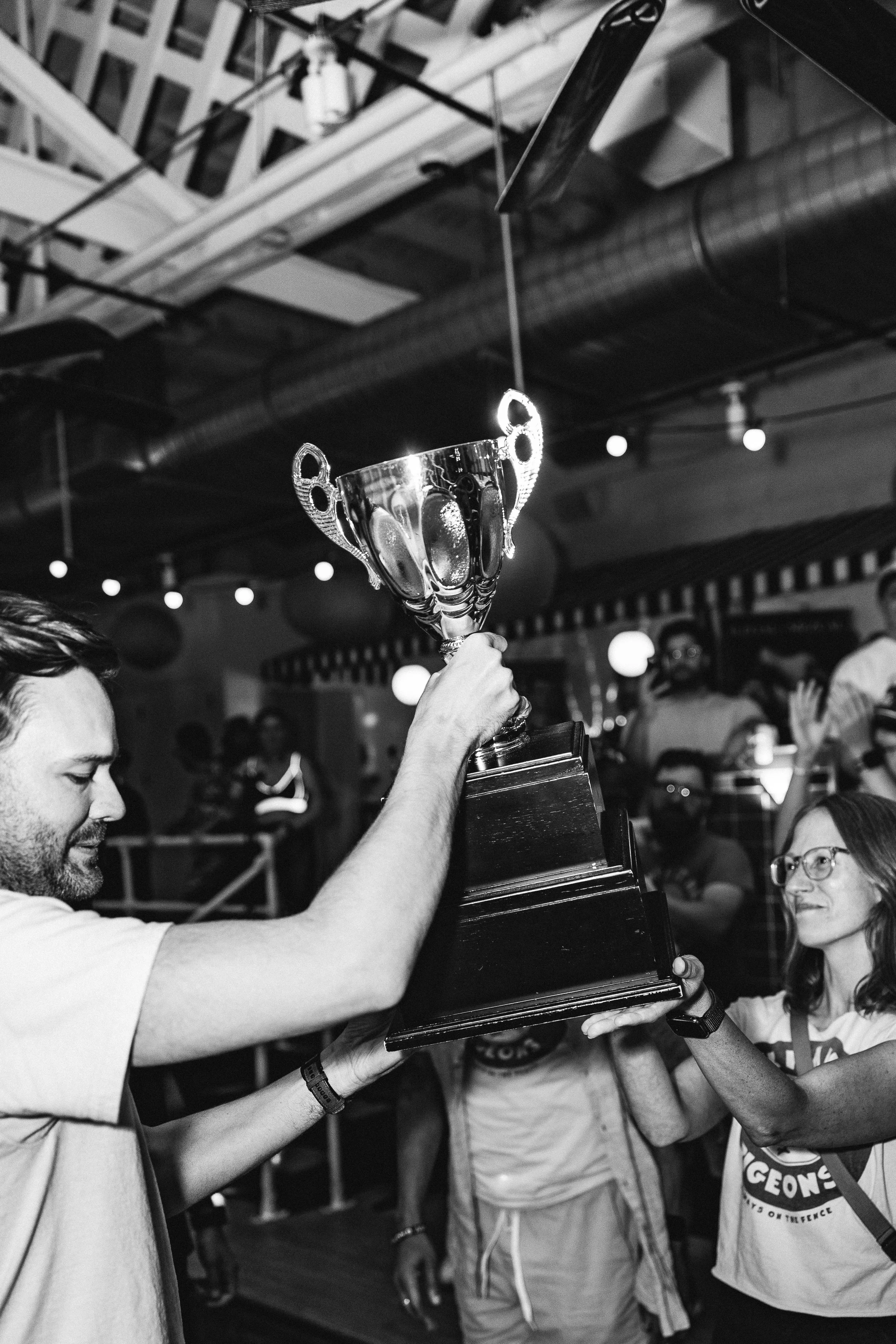 A man and a woman celebrate as they hold a large trophy during a party, with other people in the background, in a lively indoor setting.