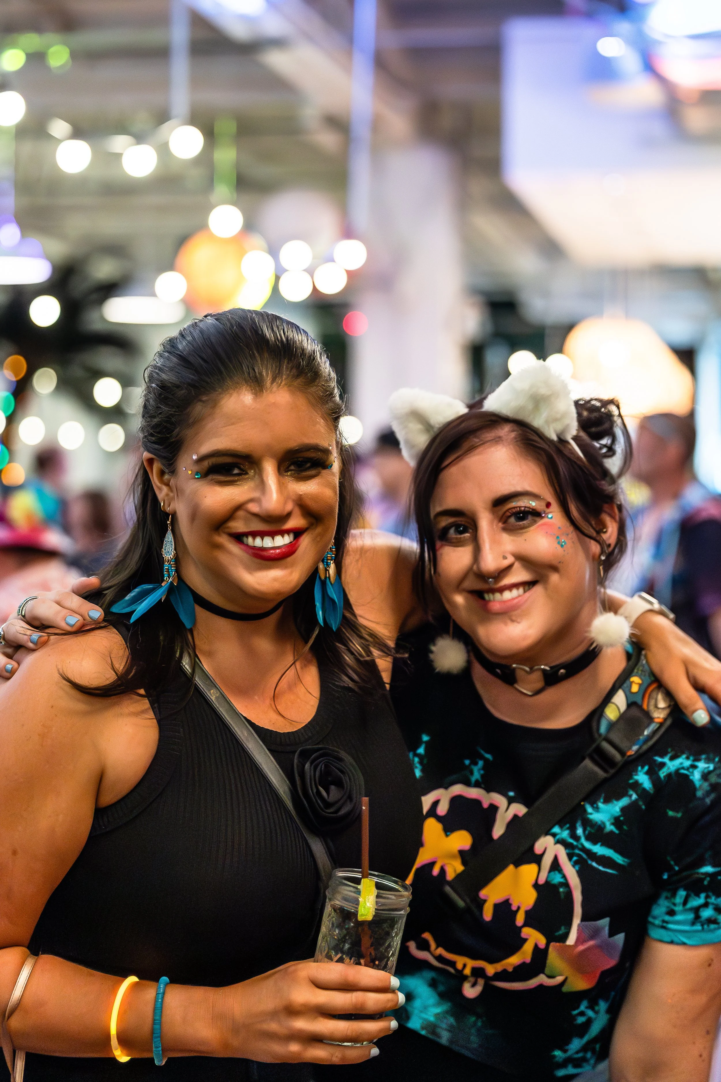 Two women smiling at a lively party or festival, with colorful lights and decorations in the background. One woman is wearing black with feather earrings and the other has white cat ears and face glitter.