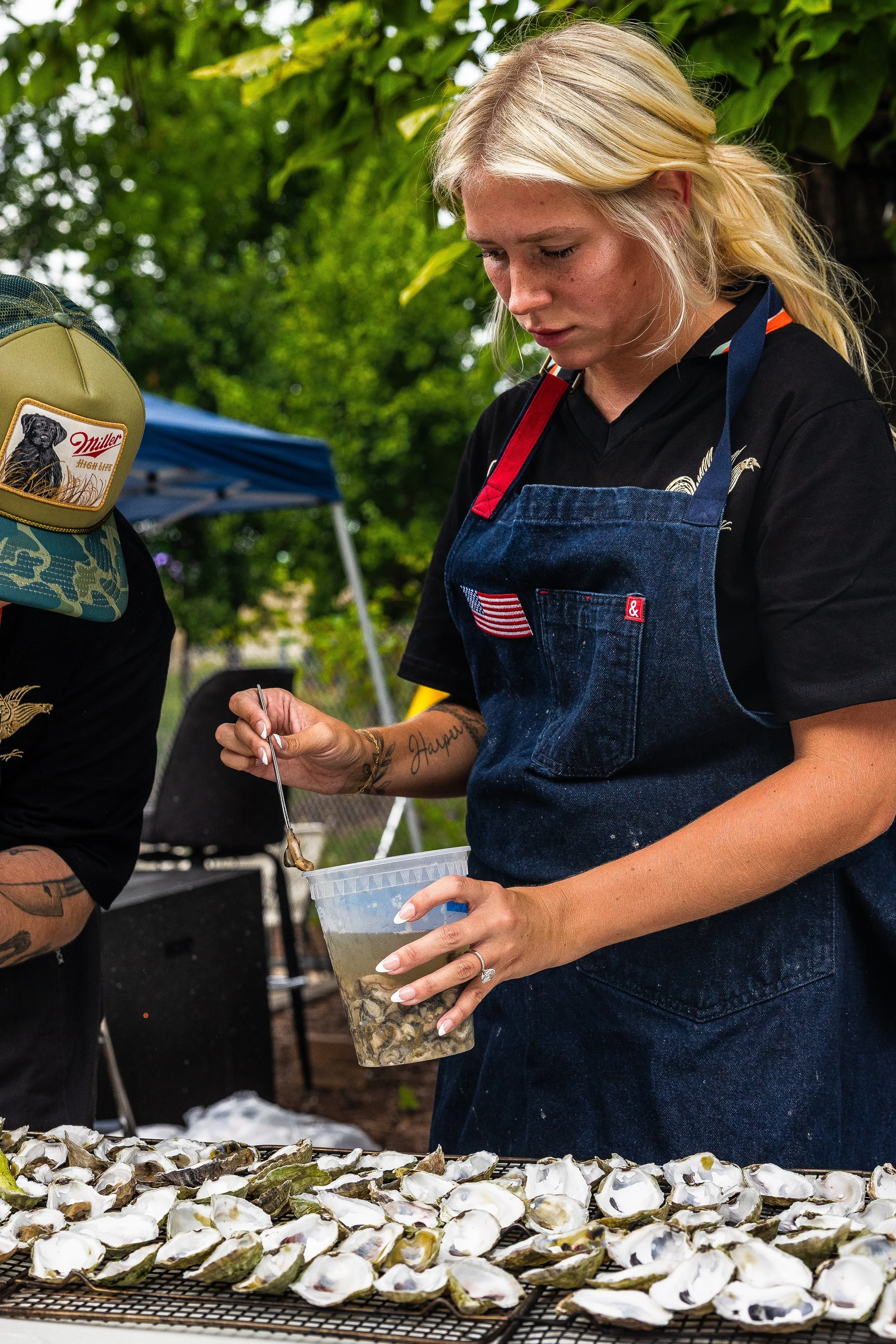 A woman shucking oysters outdoors, wearing a black shirt and denim apron, with a tray of oysters in front of her.