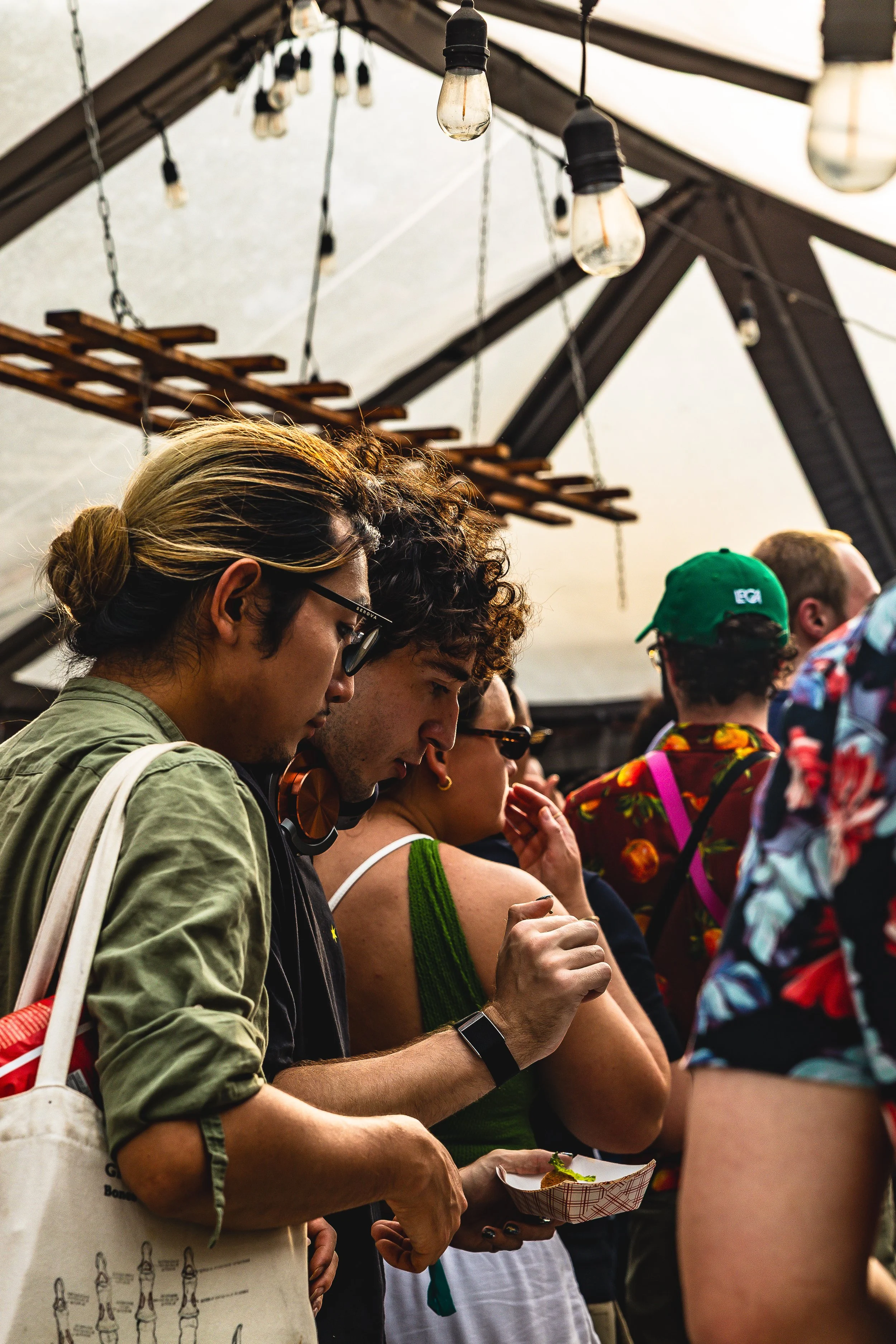 People standing in line at an indoor event under a ceiling with hanging light bulbs.