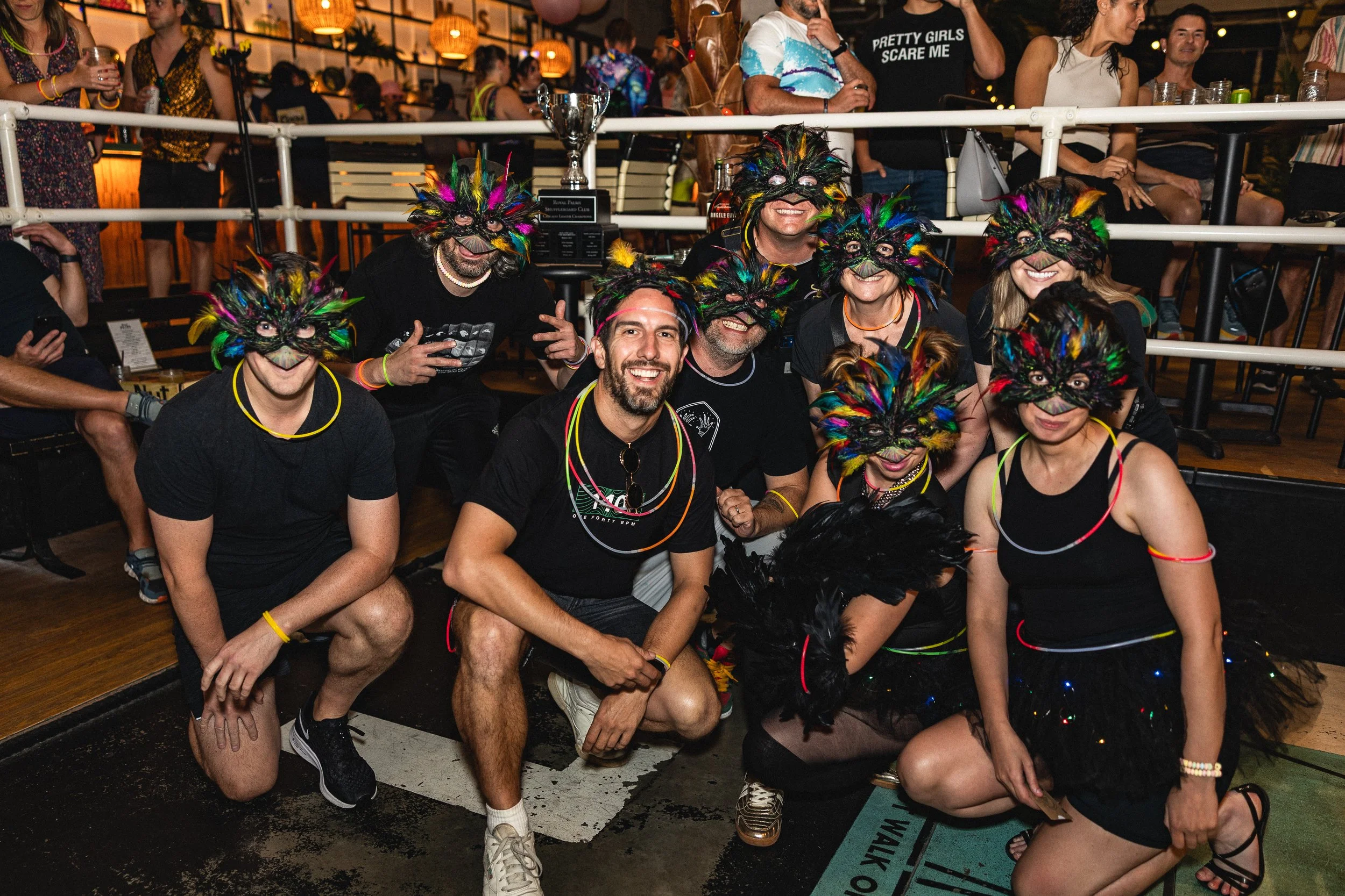 A group of people wearing colorful animal masks and necklaces, smiling and posing together at a lively indoor event with other attendees in the background.