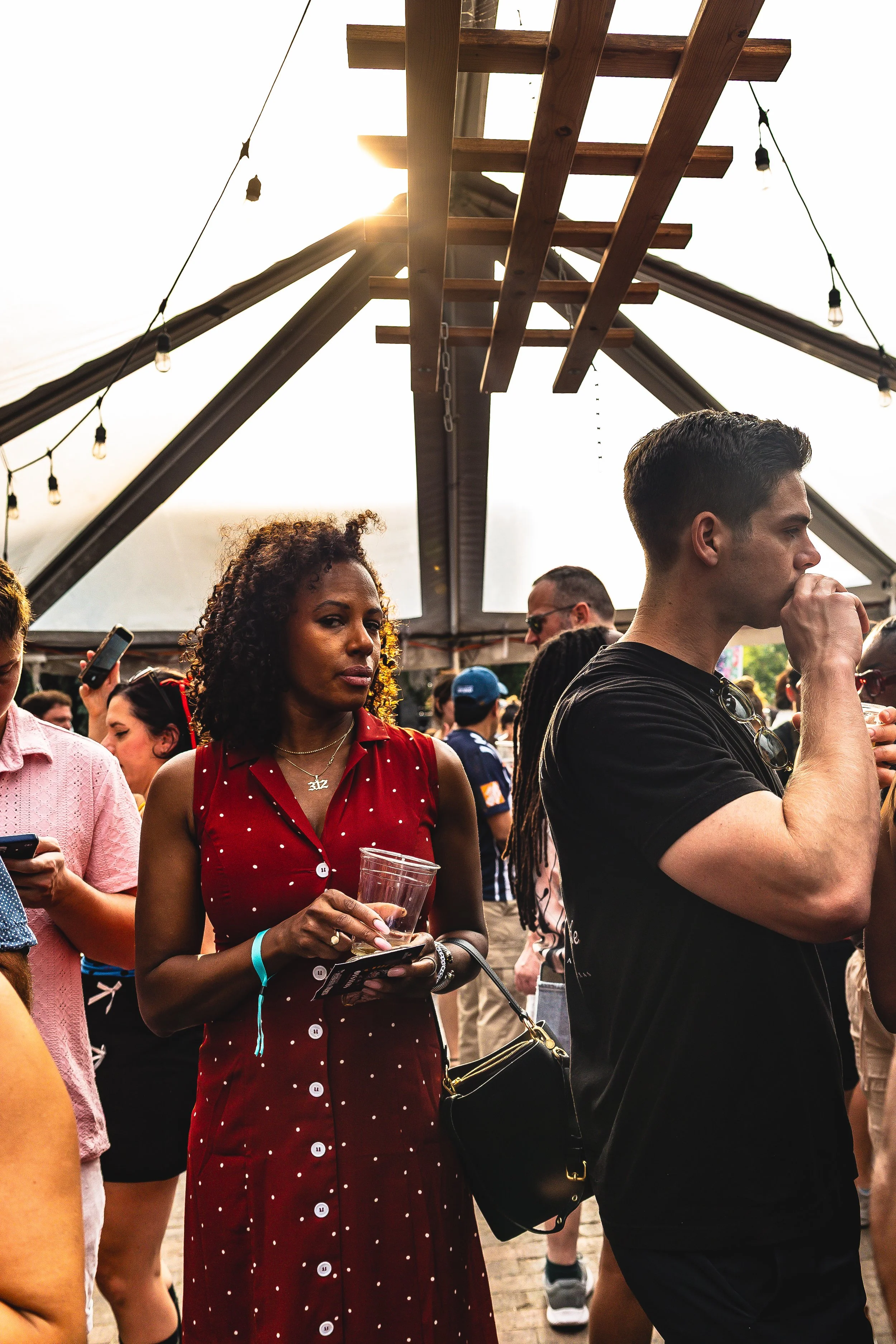 People gathered at an outdoor event, with a woman in a red polka-dot dress holding a drink in the foreground, and a man in a black shirt touching his face nearby, under a tent with string lights.