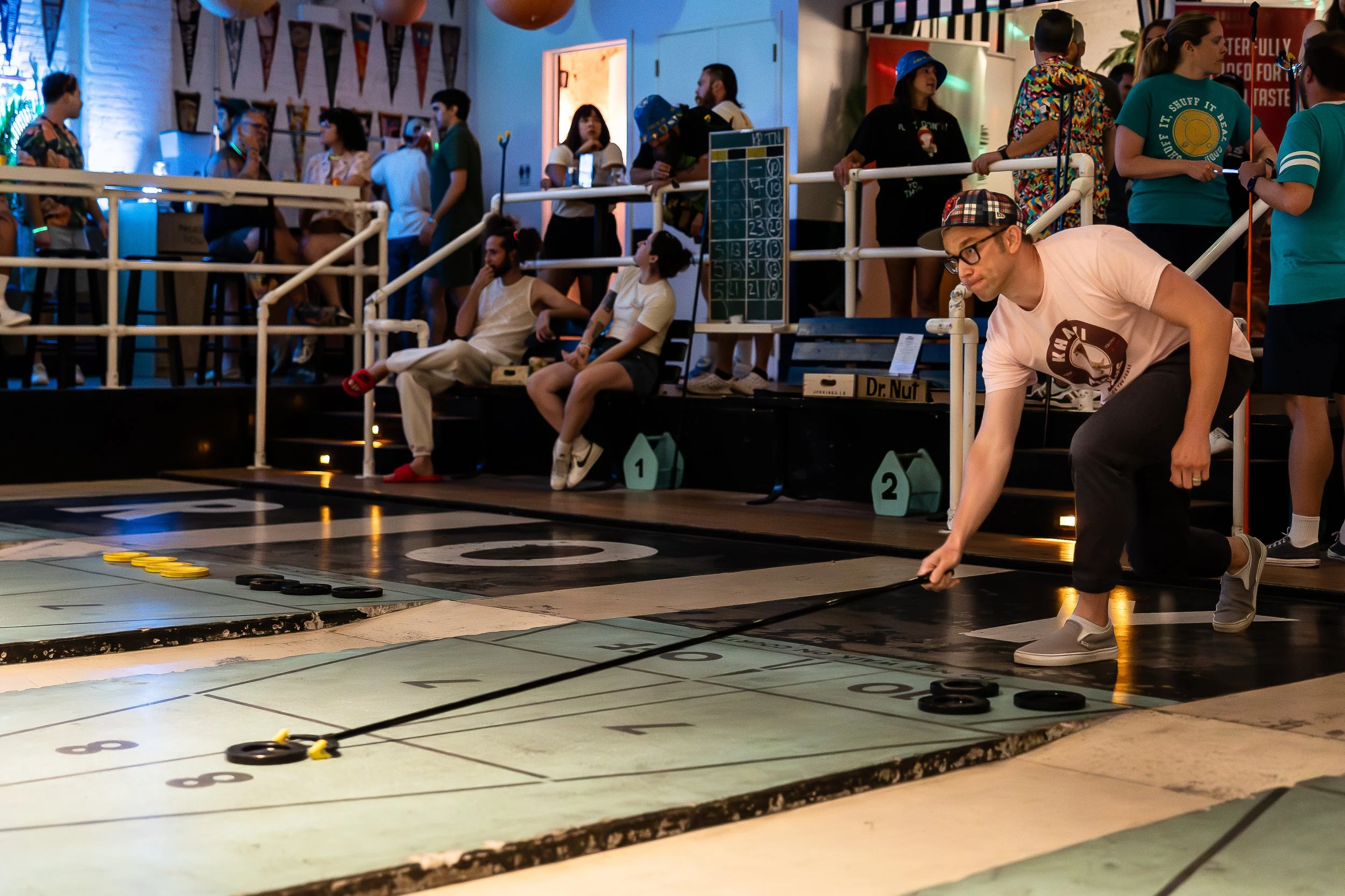 A man playing shuffleboard on an indoor court while spectators watch from benches and standing areas in the background.