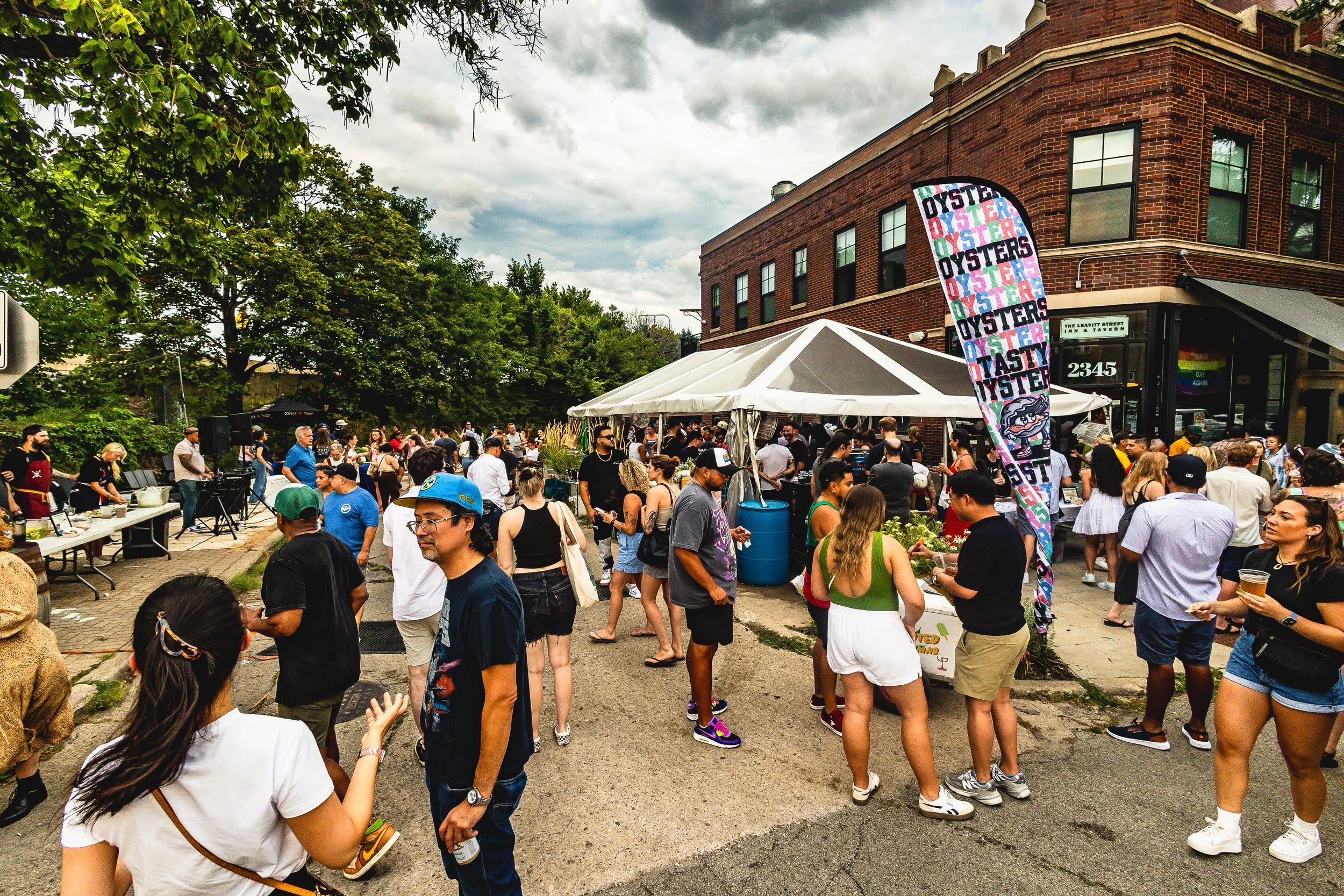 A street festival with a crowd of people, tents, and food vendors, including a banner promoting oysters. The sky is cloudy, and there are brick buildings and green trees in the background.