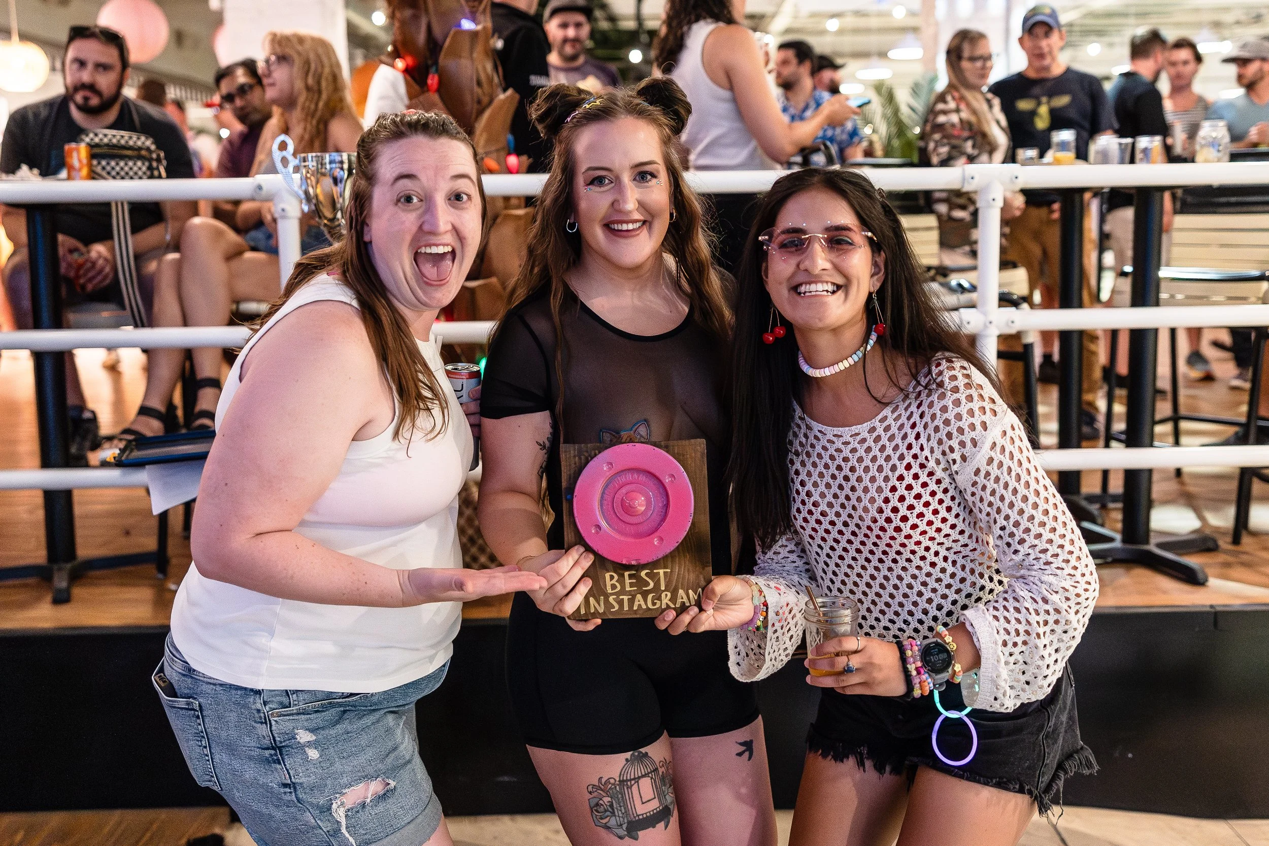 Three women smiling and holding a pink donut-shaped award that says "Best Instagram" at a party or celebration with other people in the background.