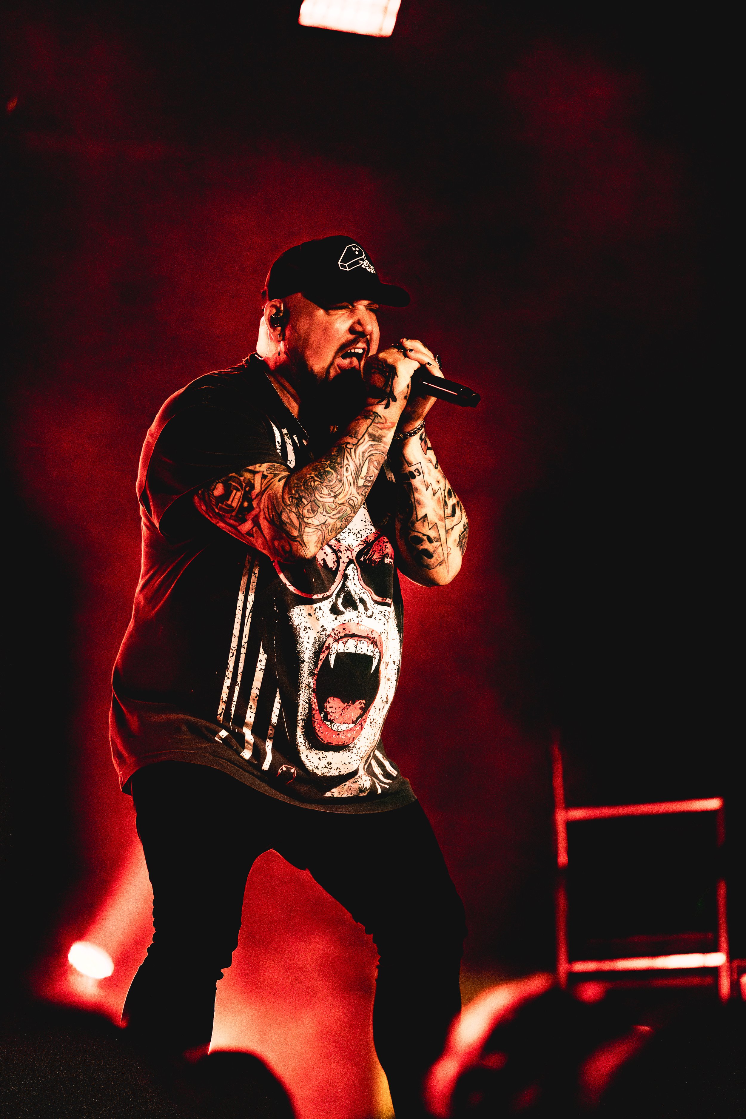 Atreyu performing passionately on stage, wearing a black cap and a graphic T-shirt with a screaming face design, holding a microphone with red stage lighting in the background.