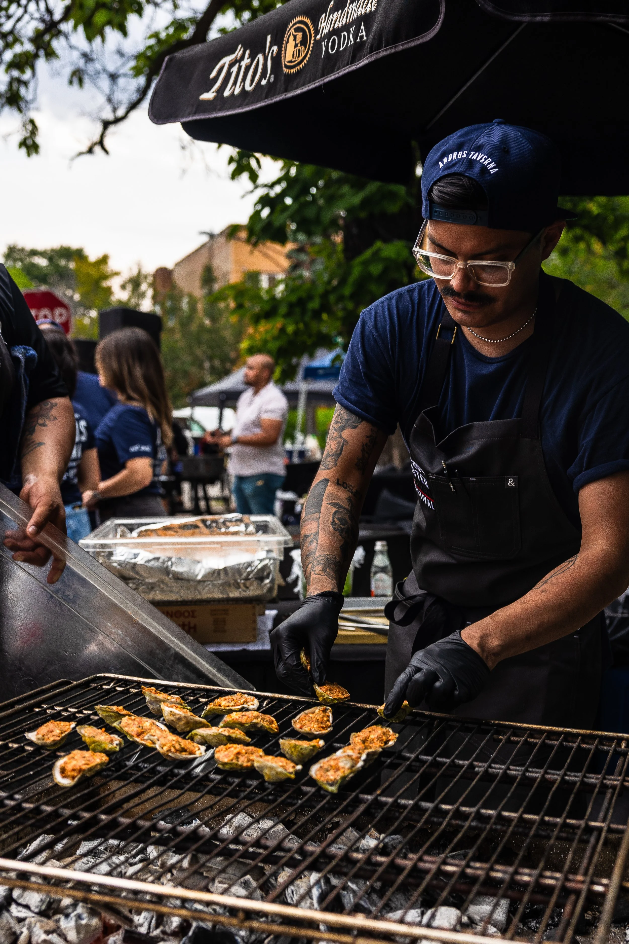 A man wearing glasses, a blue cap, and black gloves is grilling stuffed jalapeño peppers on a barbecue grill at an outdoor event. Other people are visible in the background.