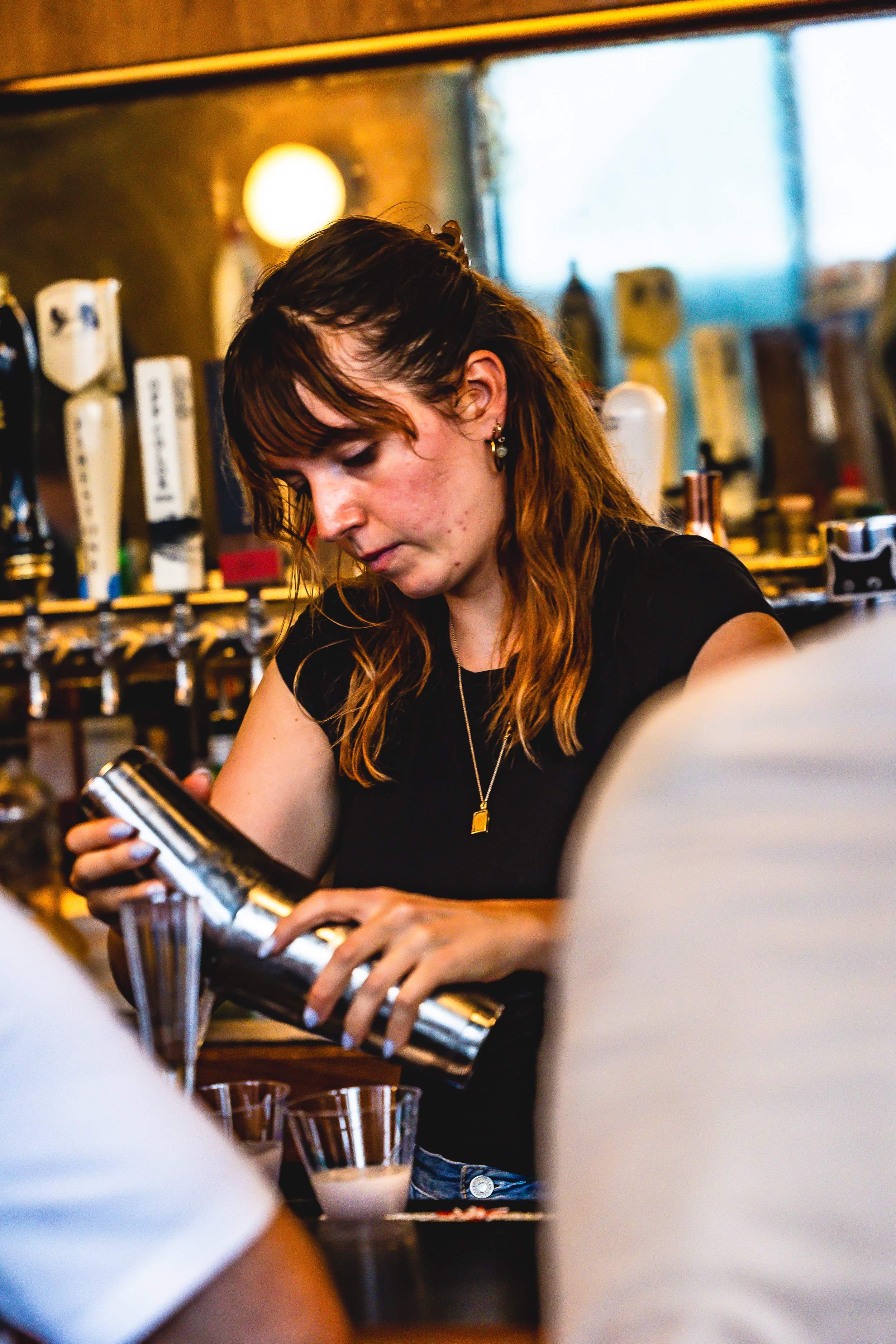 A woman with shoulder-length brown hair, wearing a black shirt and jewelry, pours a drink from a metal shaker into glasses at a bar.