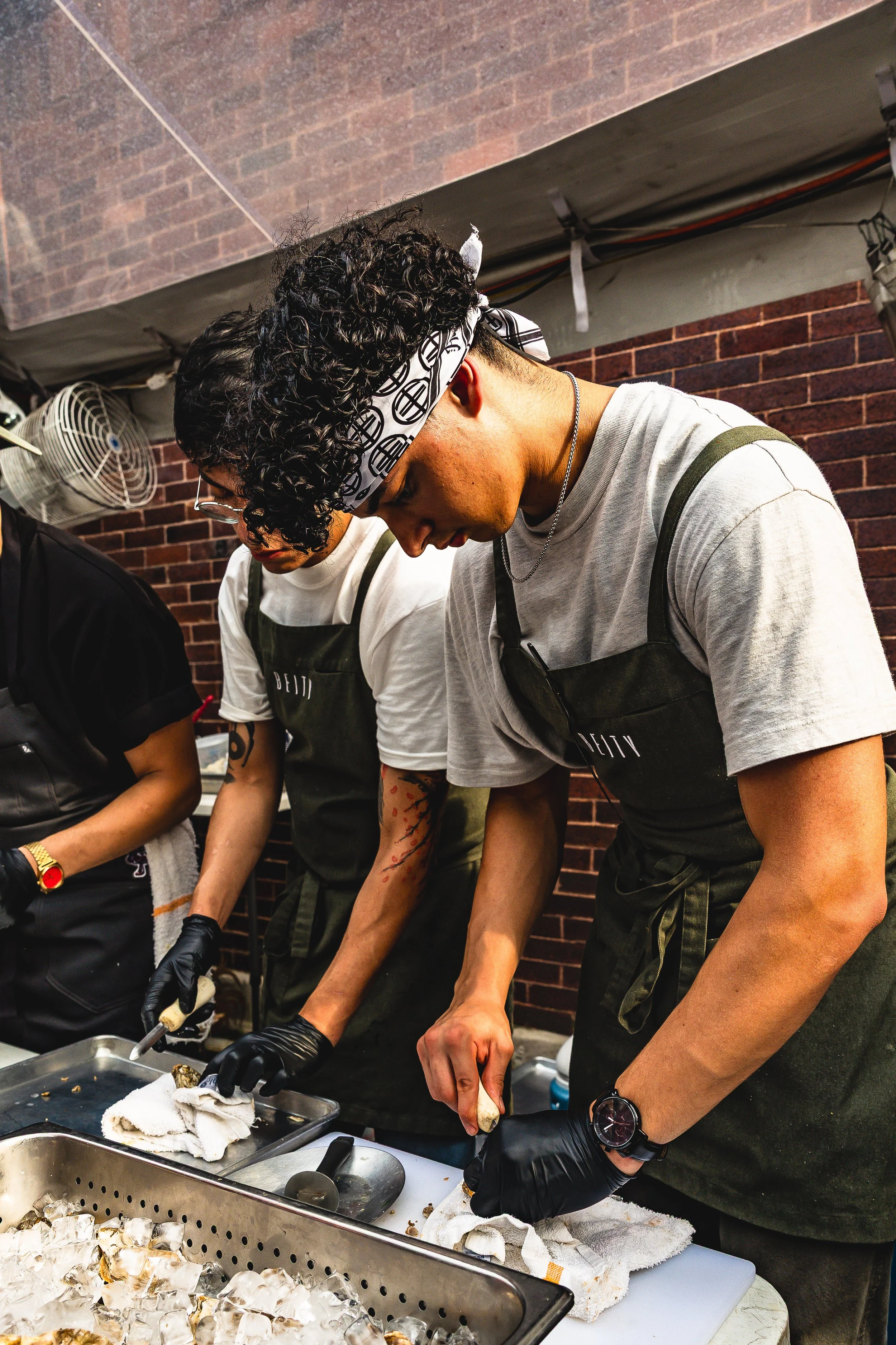 Two people wearing aprons and black gloves are shucking oysters at a table, with a brick wall in the background.
