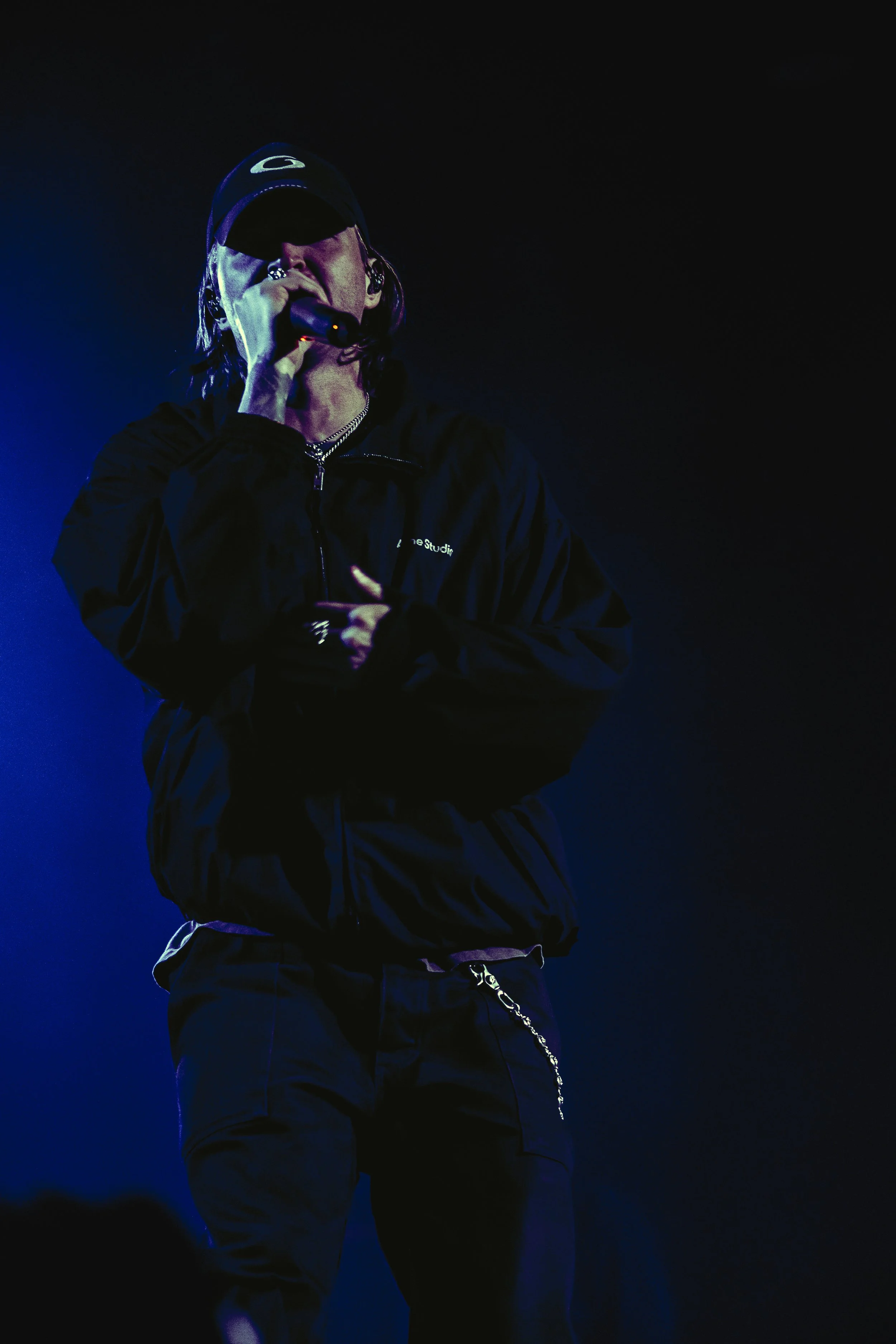 A male singer performing on stage, wearing a black jacket, black pants, a cap, and holding a microphone.